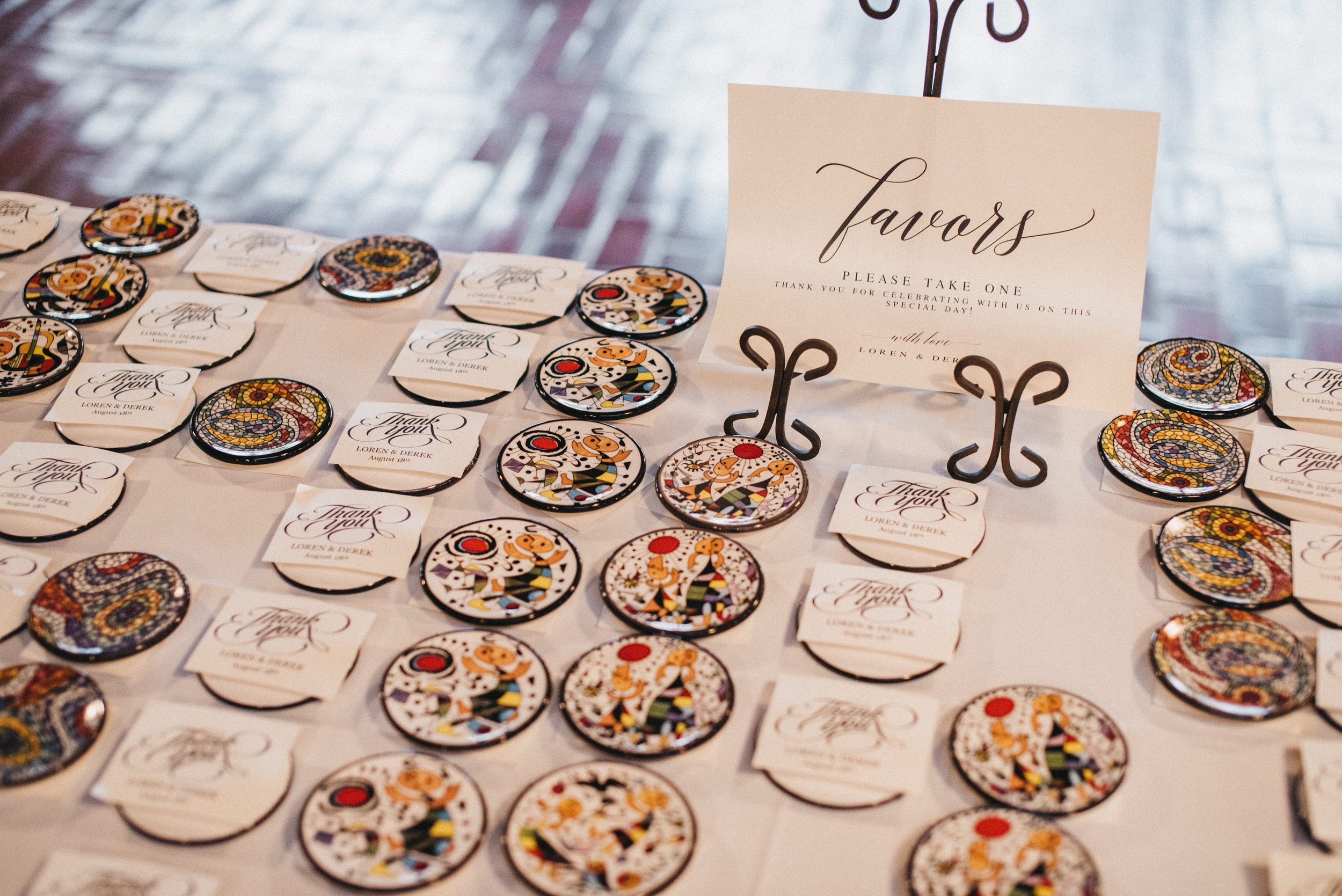 Decorative wedding favors with colorful circular badges and matching thank you notes on a table, with a sign requesting guests to take one.