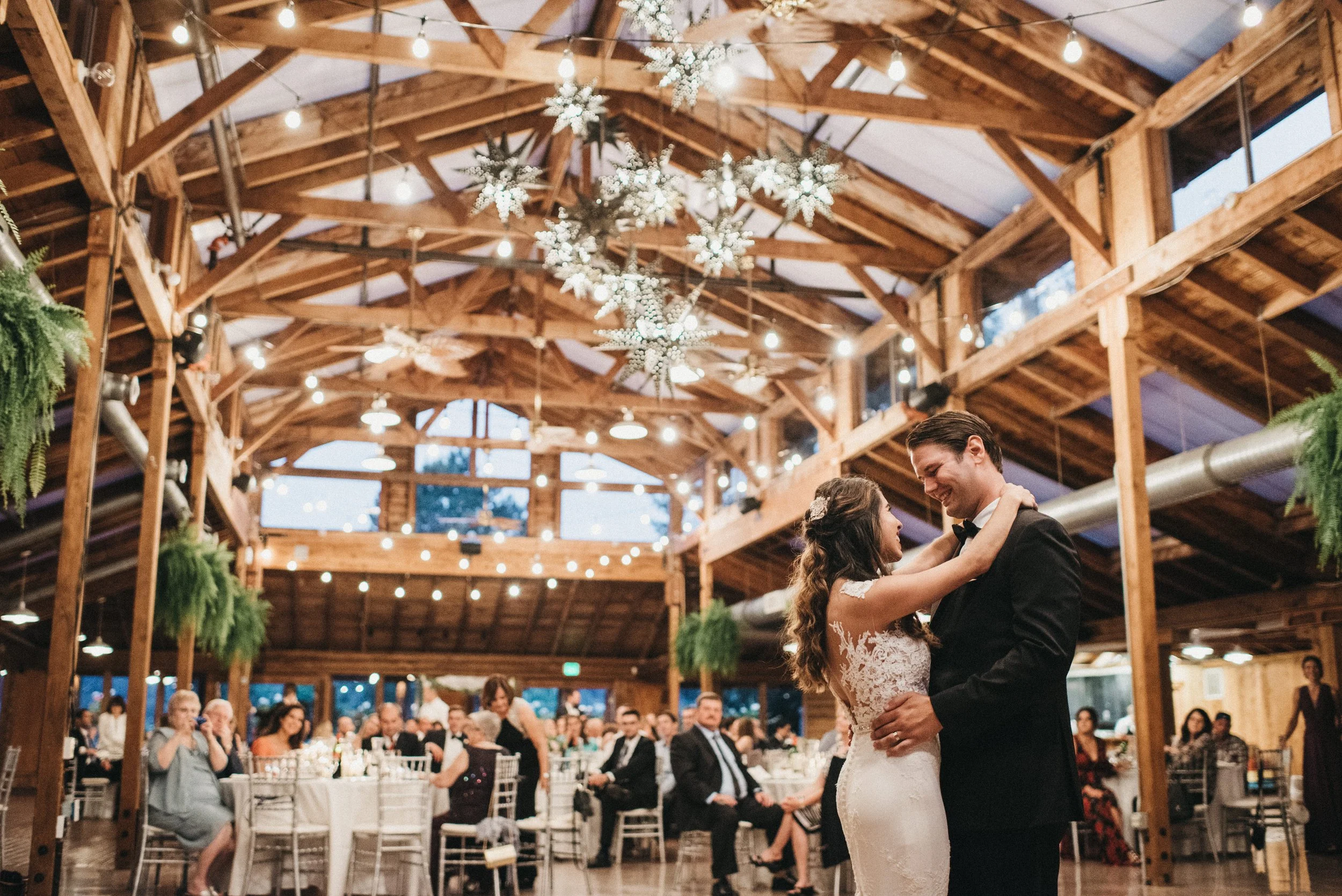 Bride and groom dancing at their wedding reception in a rustic venue with wooden beams and hanging greenery.