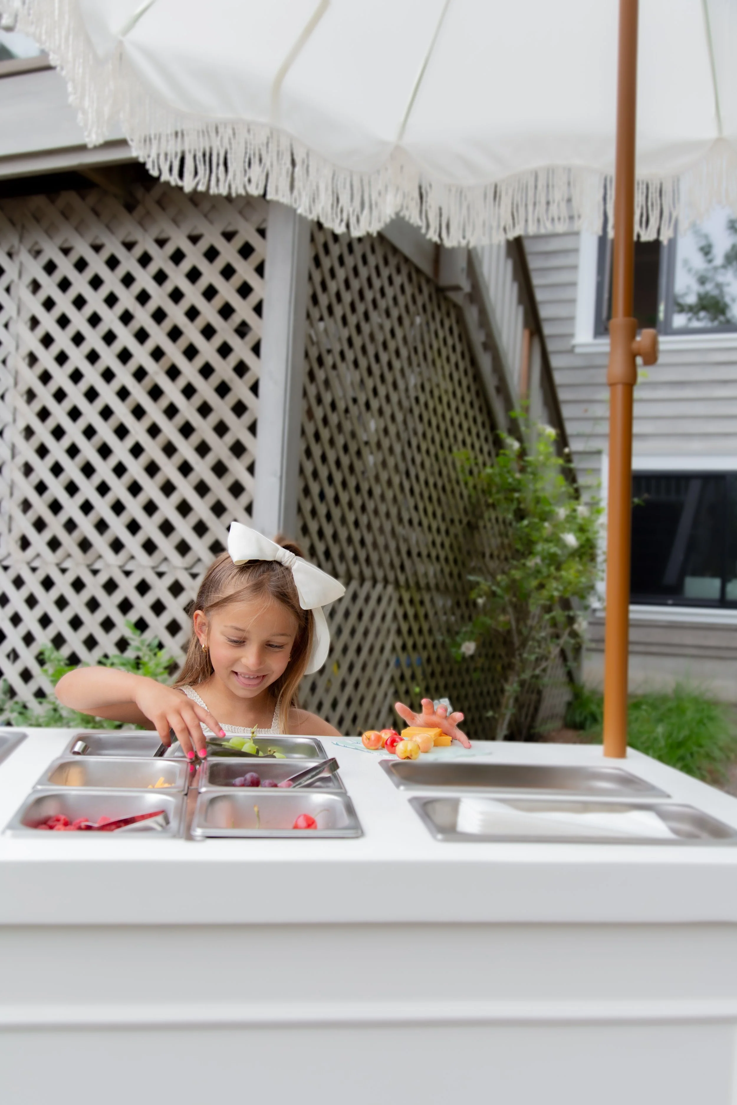 A young girl with a white bow in her hair playing at a toy ice cream shop set outdoors, with a lattice fence, a hot dog umbrella, and house window in the background.