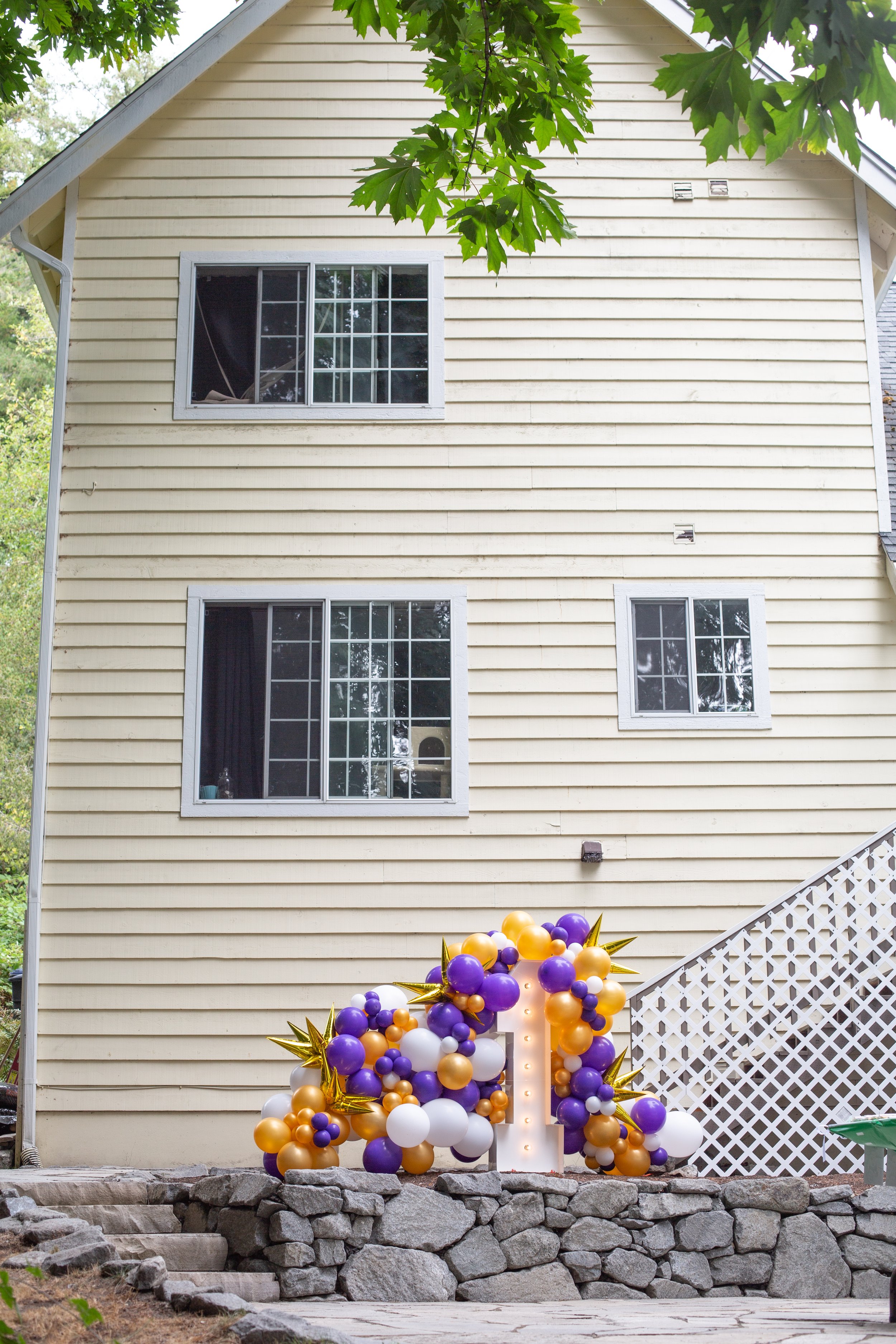 A house with beige horizontal siding, three windows, and a decorated white letter U made of balloons and lights, with purple, gold, and white balloons, some gold star-shaped balloons, and a stone foundation.