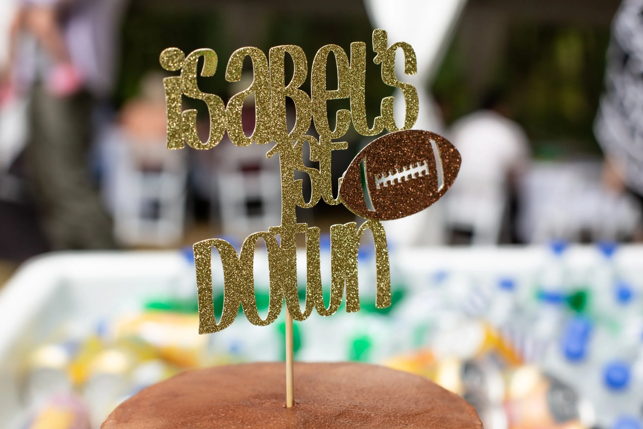 Gold glittery cake topper with a football design and the words 'SABRE'S 50th BIRTHDAY' on a chocolate cake, with blurred people and decorations in the background.