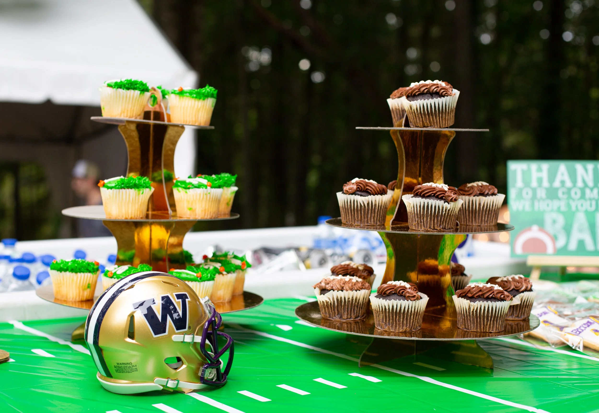 Cupcakes decorated with green icing and sprinkles, and chocolate cupcakes with swirled frosting, displayed on a multi-tiered gold cupcake stand on a table with green and white striped tablecloth, football field theme, possibly at an outdoor tailgate 