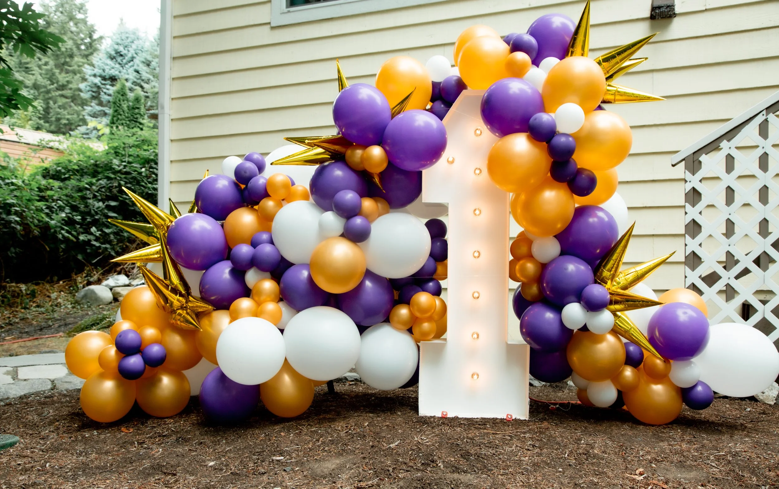 Decorative balloon arrangement with purple, gold, white, and orange balloons, surrounding a large white number one with lights, for a first birthday celebration outside a house.