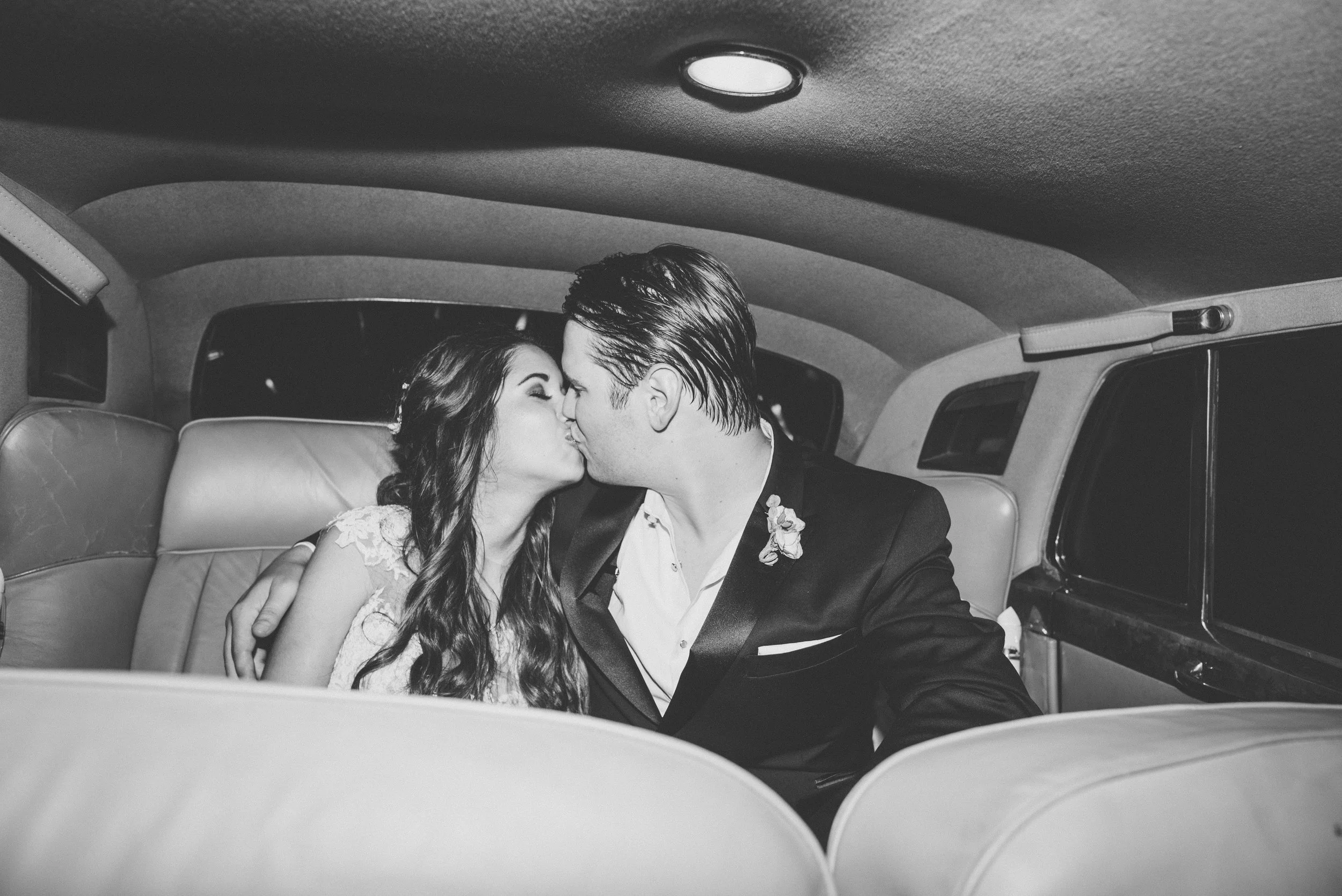 A black and white photo of a couple sharing a kiss inside a vintage car, with the woman in a lace dress and the man in a tuxedo with a boutonniere.