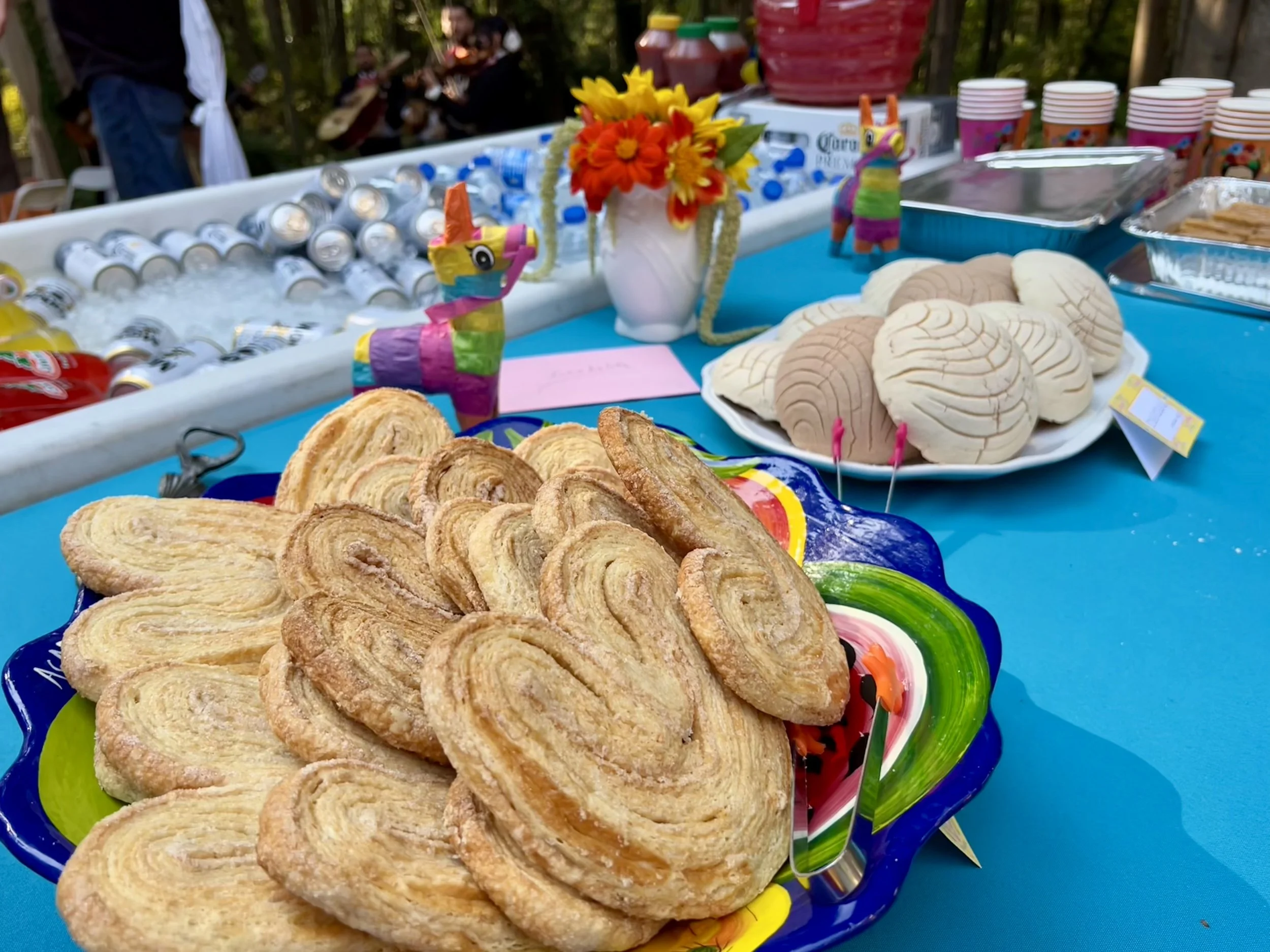 Plate of paletas, traditional Mexican frozen treats, decorated with holiday-themed picks on a blue table. In the background, there are more sweets, drinks, and decorative items, including a vase with a flower arrangement, a llama figurine, and a grou
