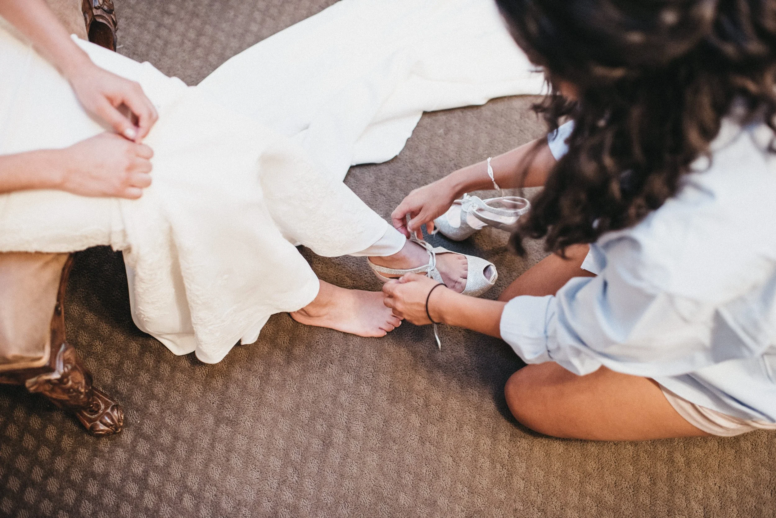 A woman in a white shirt kneels on the carpet, helping another woman in a cream dress with silver high heels.