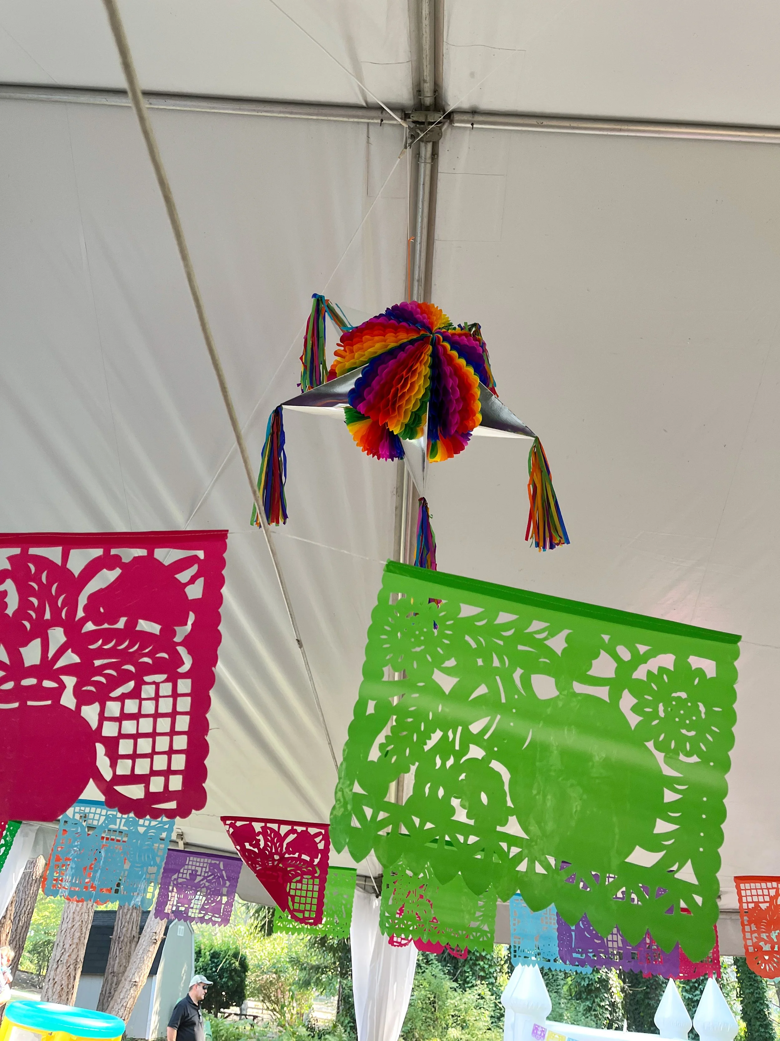 Colorful paper decorations and a rainbow paper piñata hanging inside a tent, with trees and people visible outside.