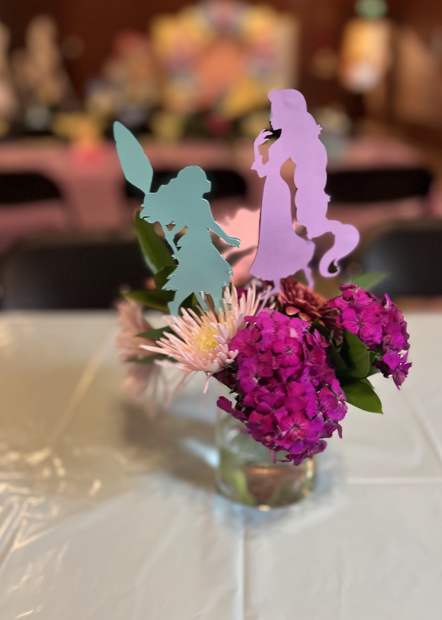 A small floral arrangement in a glass jar with pink and purple flowers, topped with paper cutouts of Tinker Bell and a mermaid from Disney.