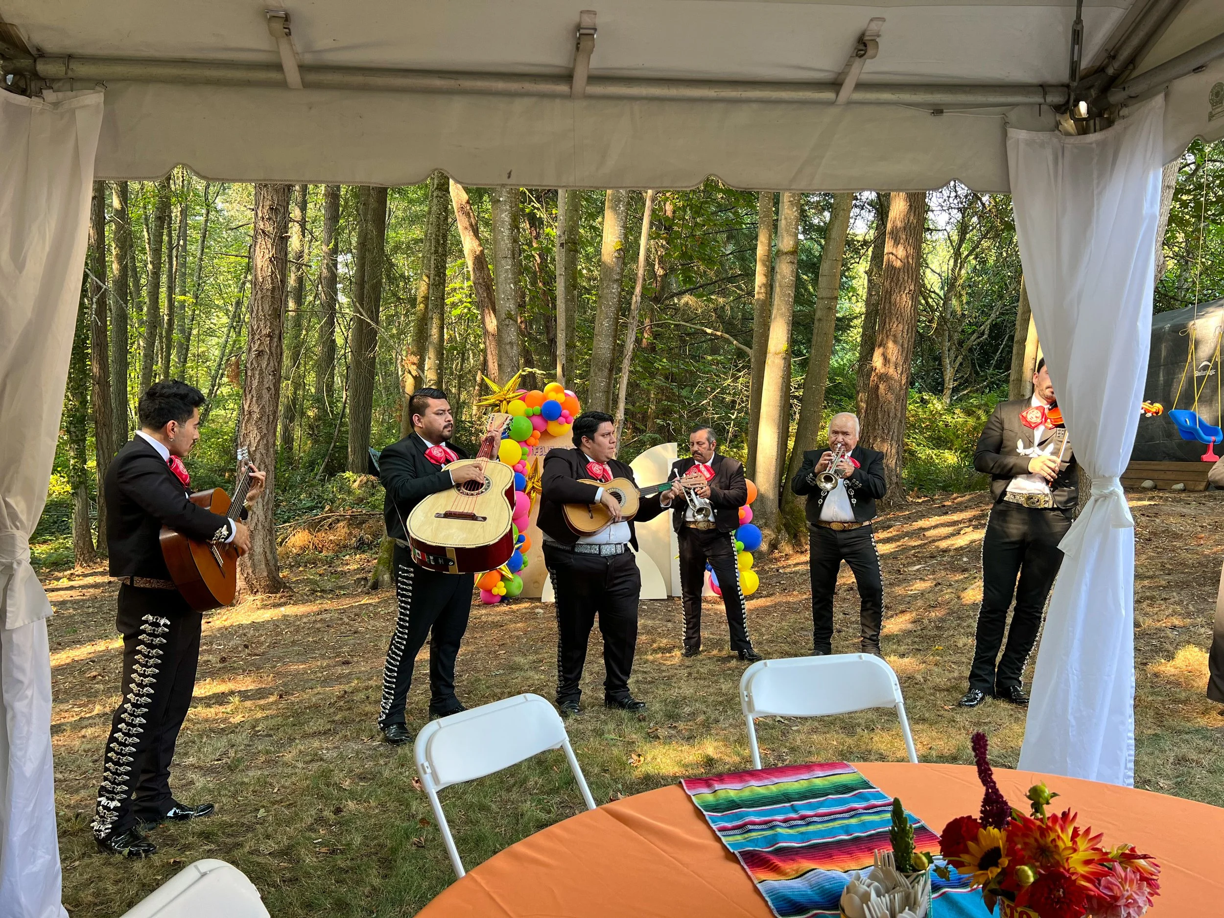 Musicians performing outdoors in a wooded area during a celebration, with colorful balloons and decorations in the background, and a table with a colorful striped cloth and flowers in the foreground.