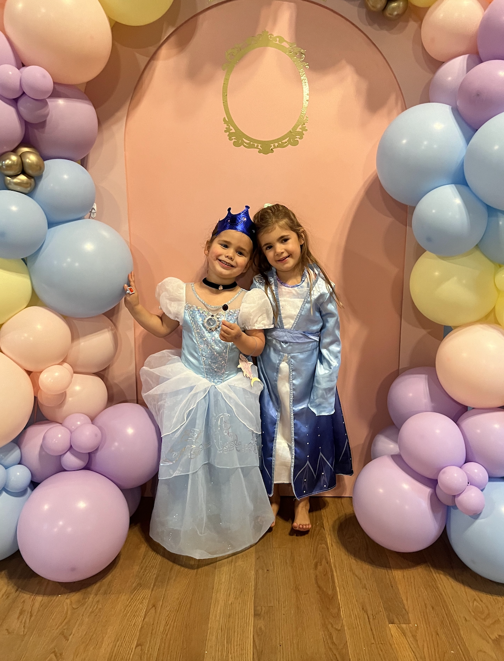 Two young girls dressed as princesses, one in a light blue gown with a crown and the other in a blue and white gown, standing together in front of a pink backdrop decorated with pastel-colored balloons.