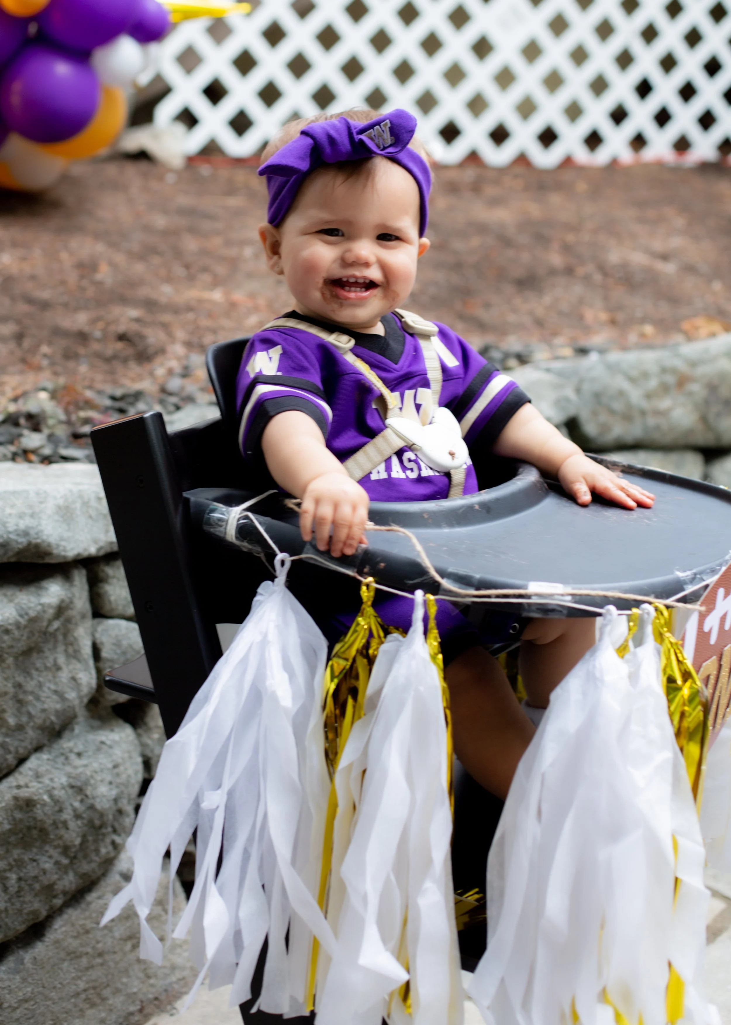 A smiling baby in a purple football jersey and matching headband sitting in a black high chair decorated with white and gold streamers, outdoors with rocks and a lattice fence in the background.