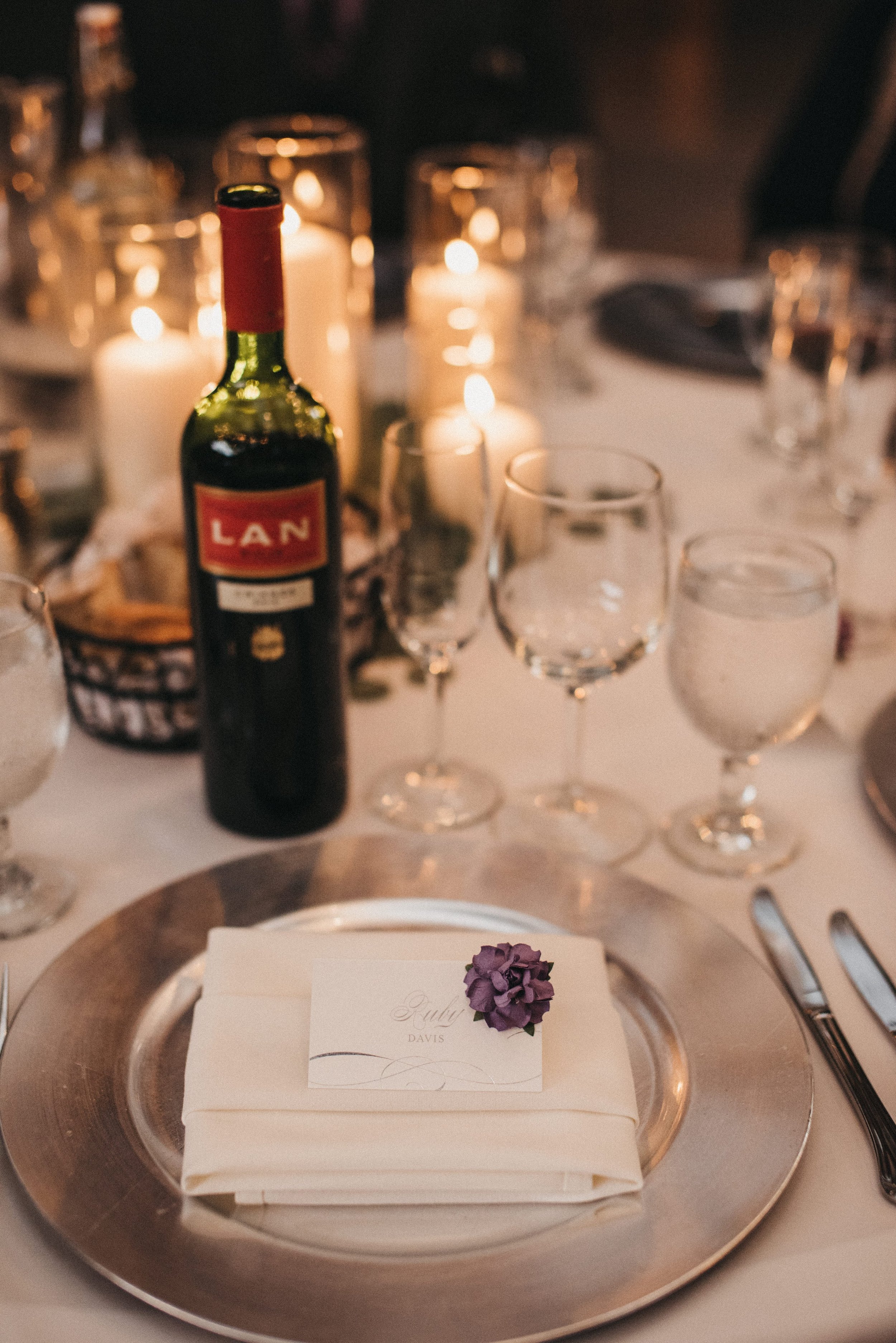 A wedding or formal dining table setting with a silver charger plate, folded napkin, place card with a purple flower, wine glass, water glass, bottle of red wine, and candles in the background.