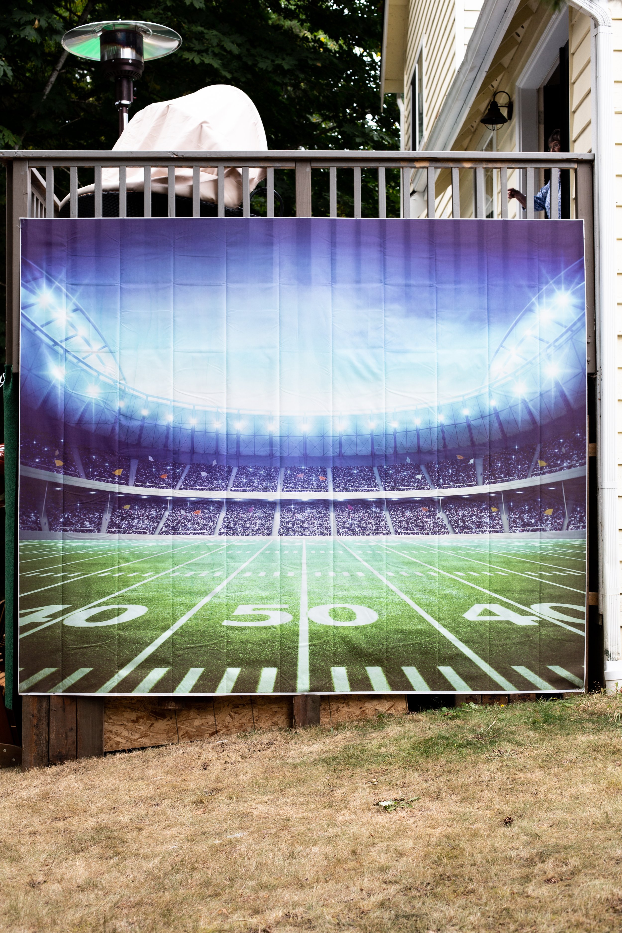 A large banner of a football field graphic mounted on the outside wall of a house, showing yard lines from 30 to 50, with a stadium crowd and bright stadium lights in the background.