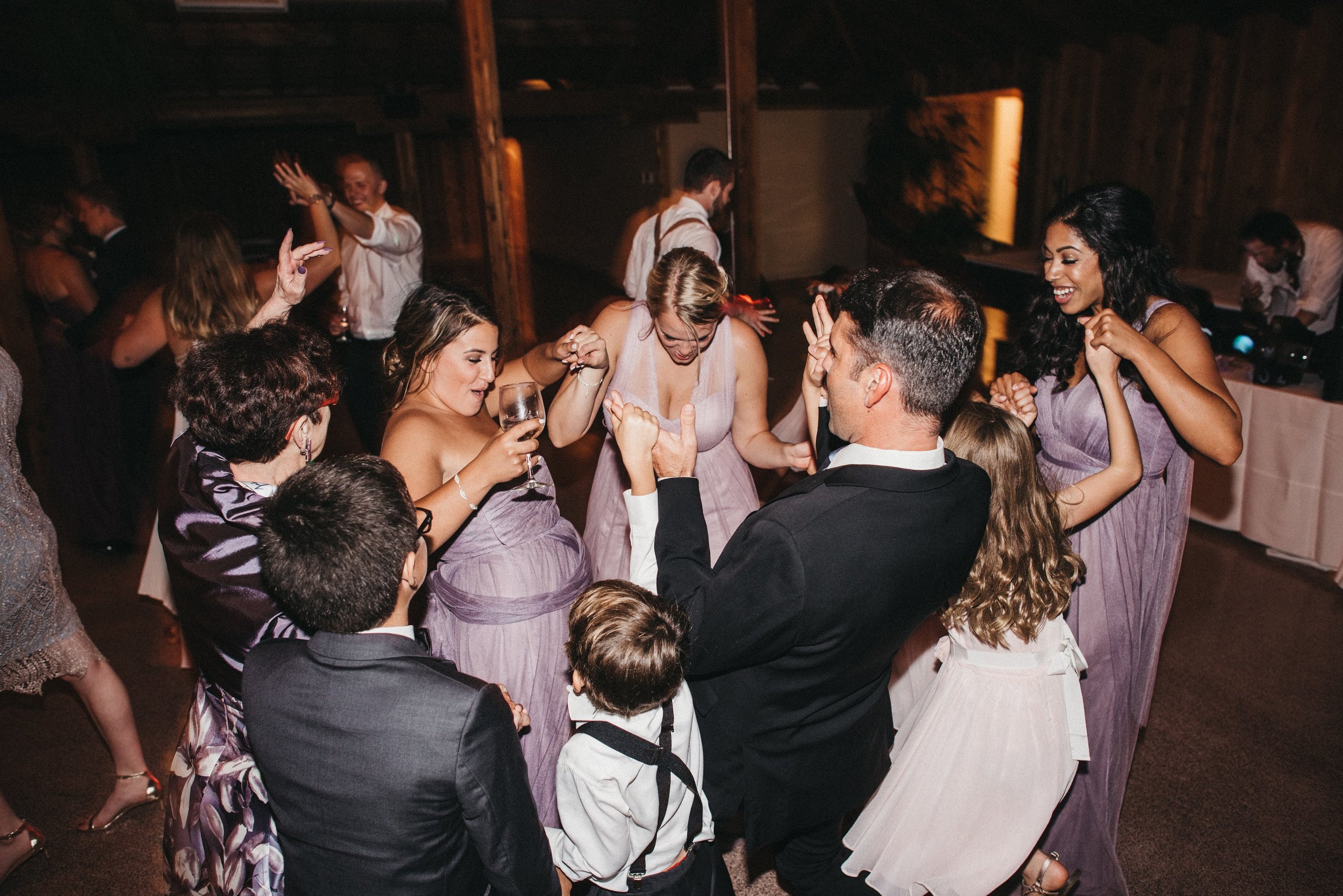 Group of people dancing and celebrating at a party or wedding reception, with women in purple dresses, men in suits, and children present.