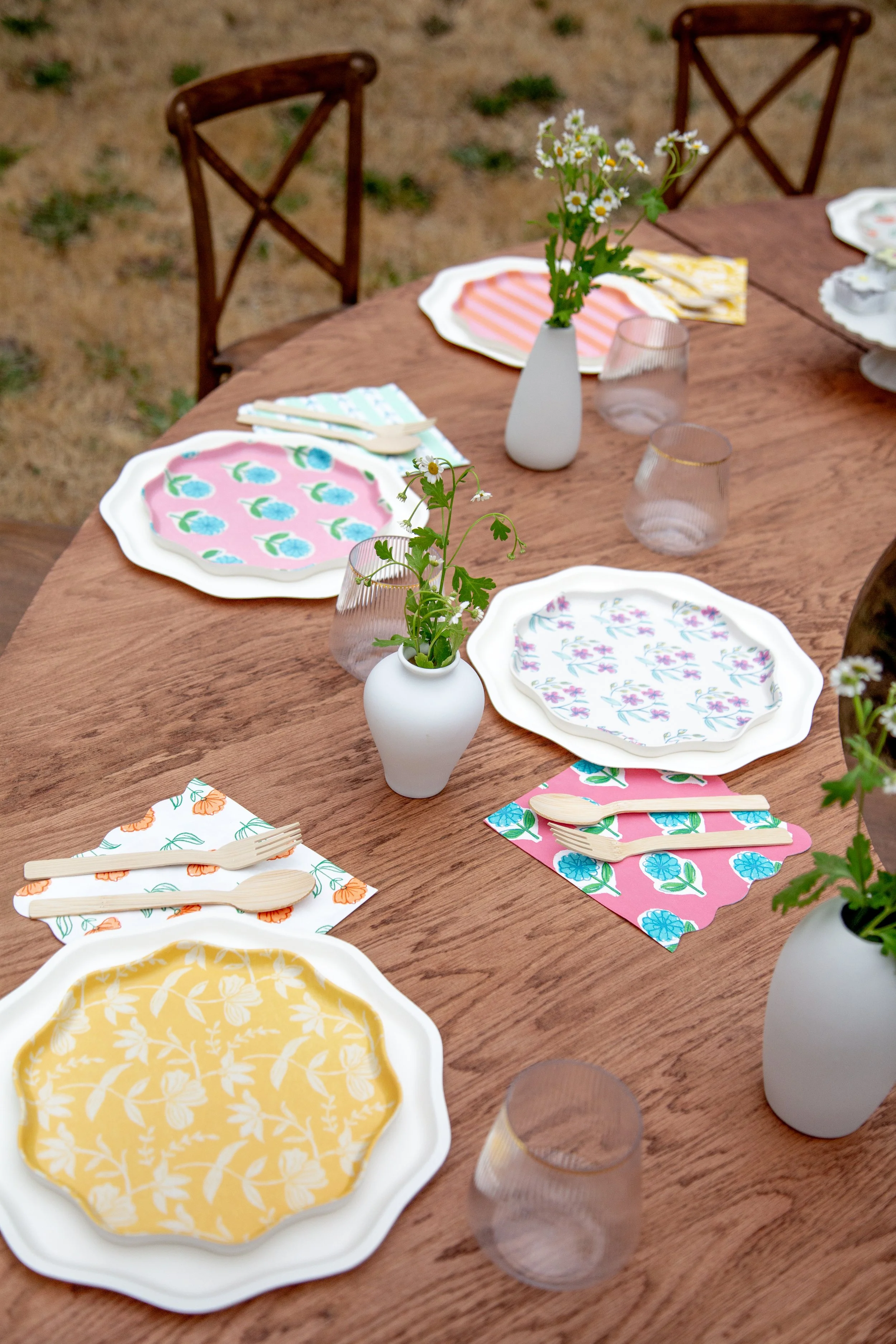 A wooden outdoor table set for a meal with colorful plates, wooden utensils, pink and yellow napkins, clear glasses, and white vases with daisies.