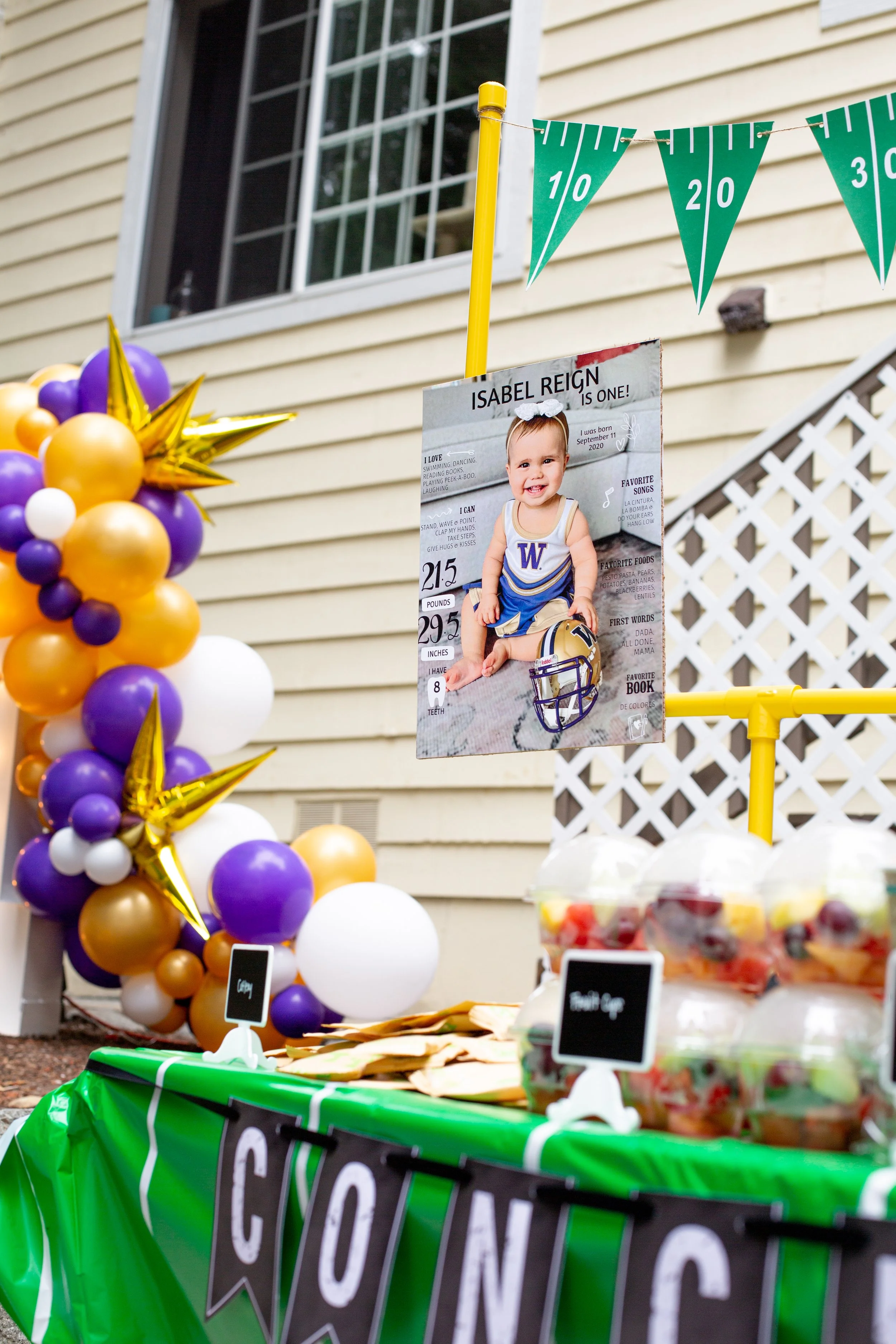 A decorated outdoor celebration for a child's first birthday, featuring purple, gold, and white balloons, a photo poster of a baby girl in a football uniform, and a table with snacks and signs.