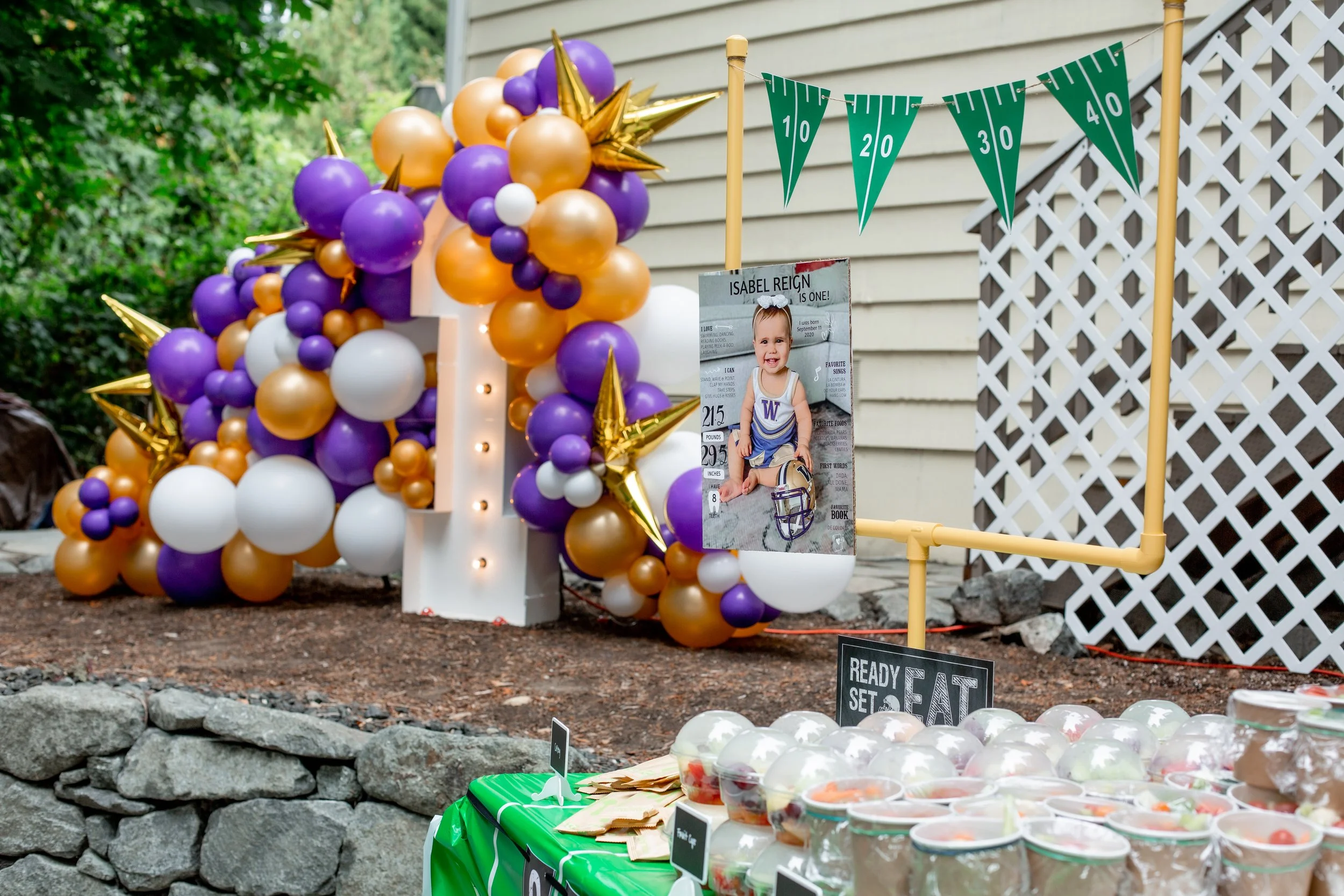 Decorative birthday celebration setup with a balloon arch in purple, white, gold, and orange, featuring a large number '1' illuminated in the center. A photo of a smiling young girl in a sports outfit with the name 'Isabel Reign' is hanging nearby. G