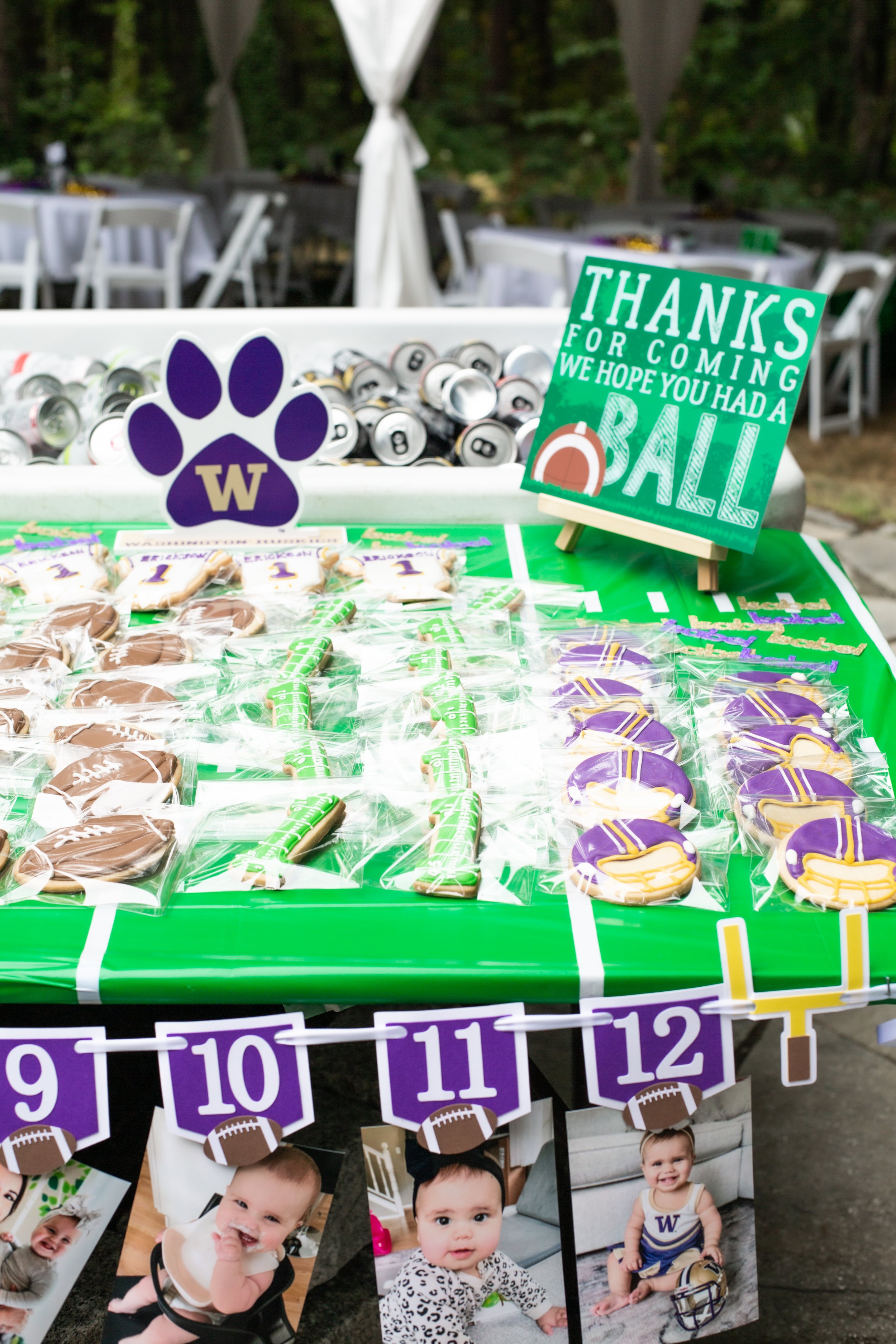 Party table decorated with football-themed cookies, a purple paw print sign with a 'W' for University of Washington, and a green sign thanking guests for coming to a football-themed event. The table is set outdoors with chairs and trees in the backgr