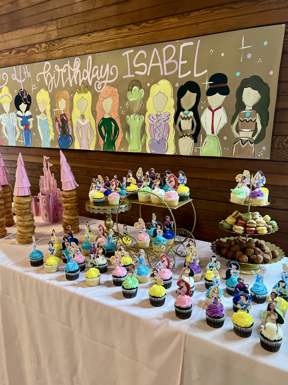 A birthday party dessert table decorated with colorful cupcakes featuring princess toppers, stacked donut towers resembling castles, and a variety of cookies and macarons. A large illustrated banner on the wood-paneled wall shows princess characters 