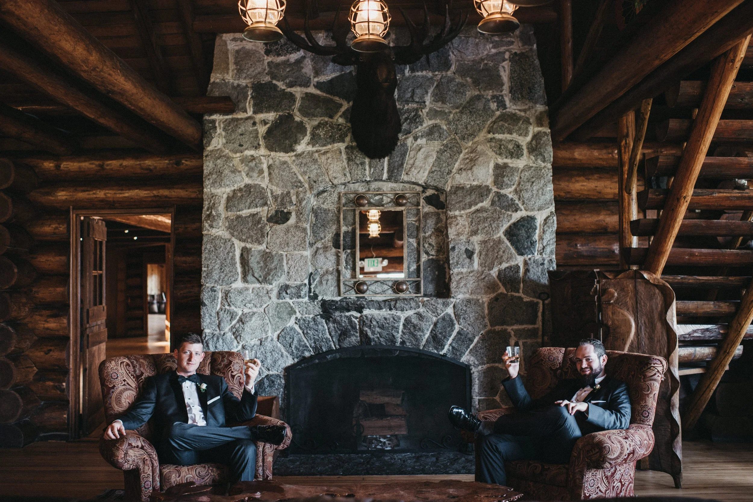 Two men in tuxedos sitting on antique armchairs, holding glasses, in front of a stone fireplace inside a log cabin, with a moose head mounted above the fireplace.