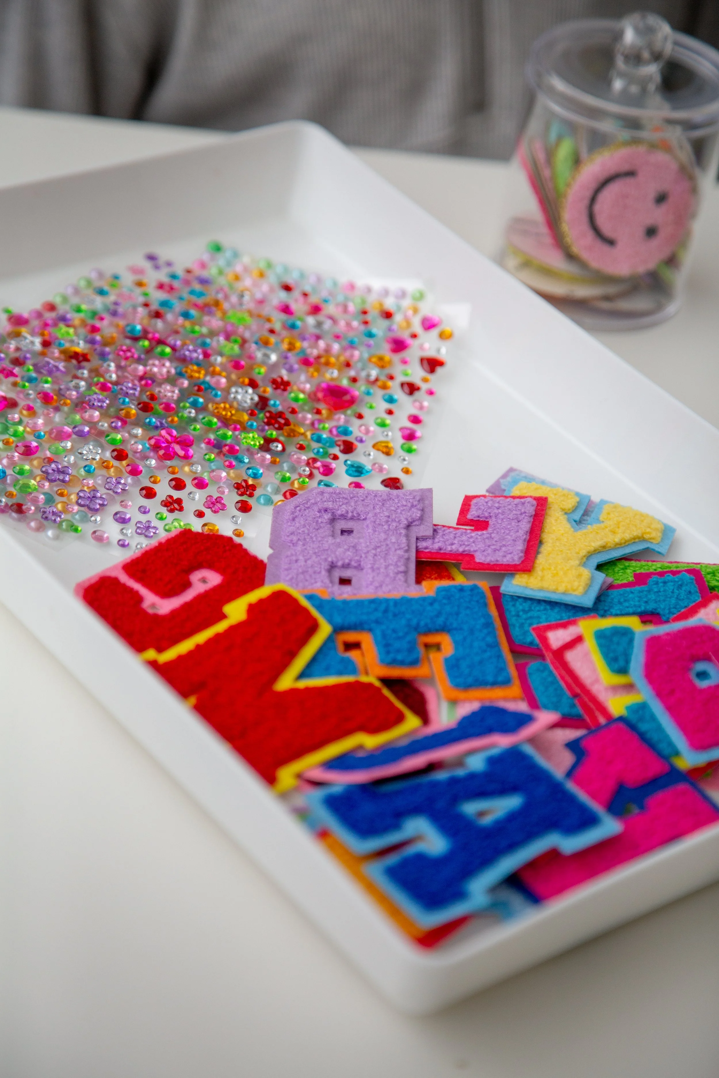 Colorful craft supplies including assorted rhinestones, felt letters, and decorative patches on a white tray.