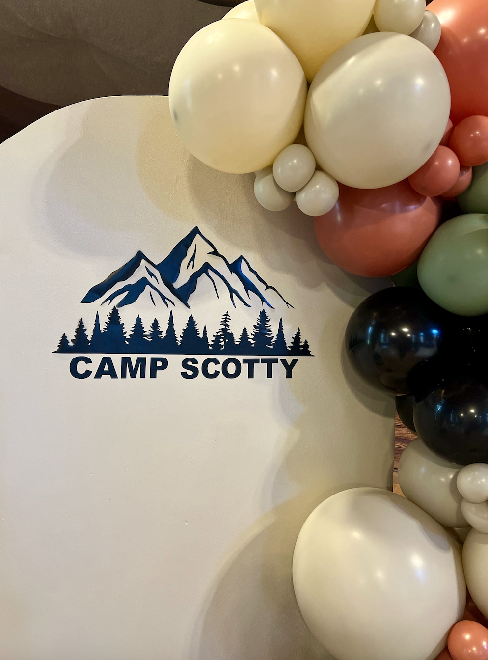 A white decorative board with a mountain and forest illustration, reading "Camp Scotty," bordered by a cluster of multicolored balloons including white, pink, black, and green.