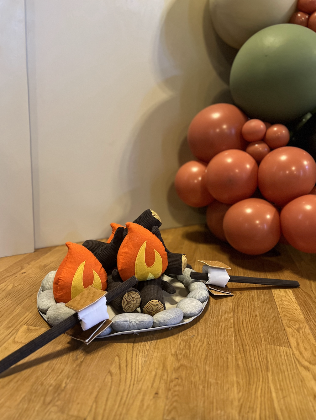 A plush toy of a black and gray creature with orange fireball plushies on its back, sitting on a gray padded ring, on a wooden floor, with a background of fabric-covered walls and decorative balloons resembling a cluster of red, green, and beige sphe