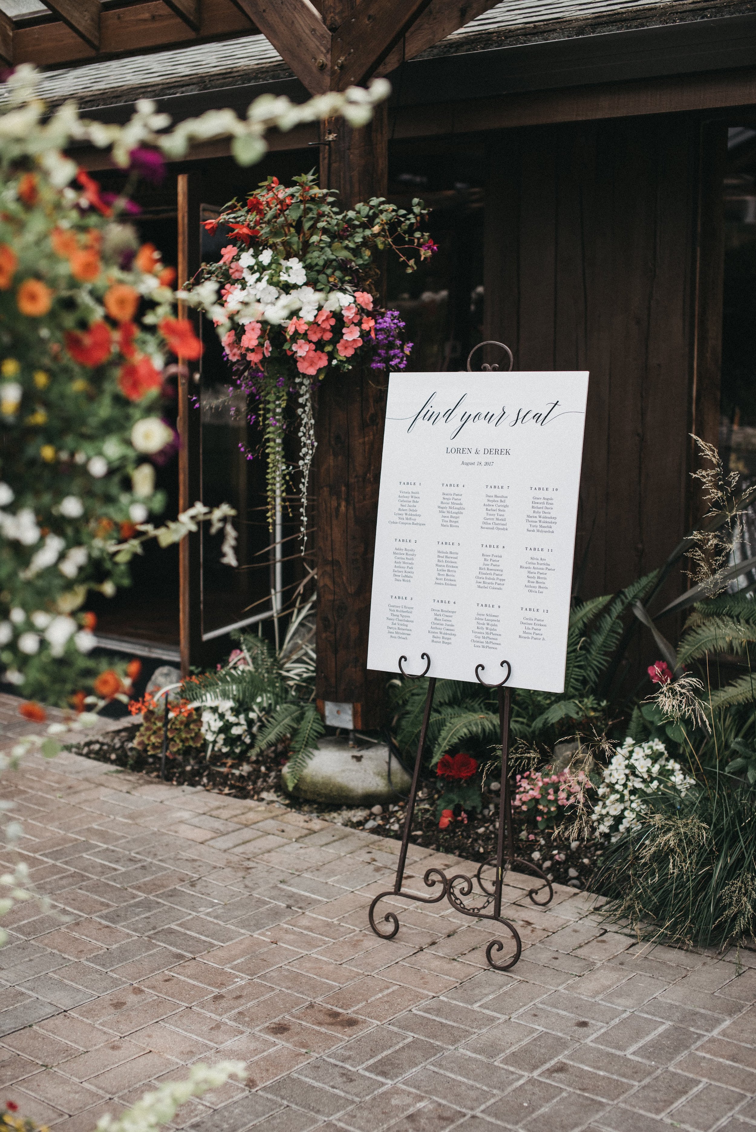 A wedding seating chart on a black metal stand with floral decorations, including pink, purple, and white flowers, outside a rustic wooden building.