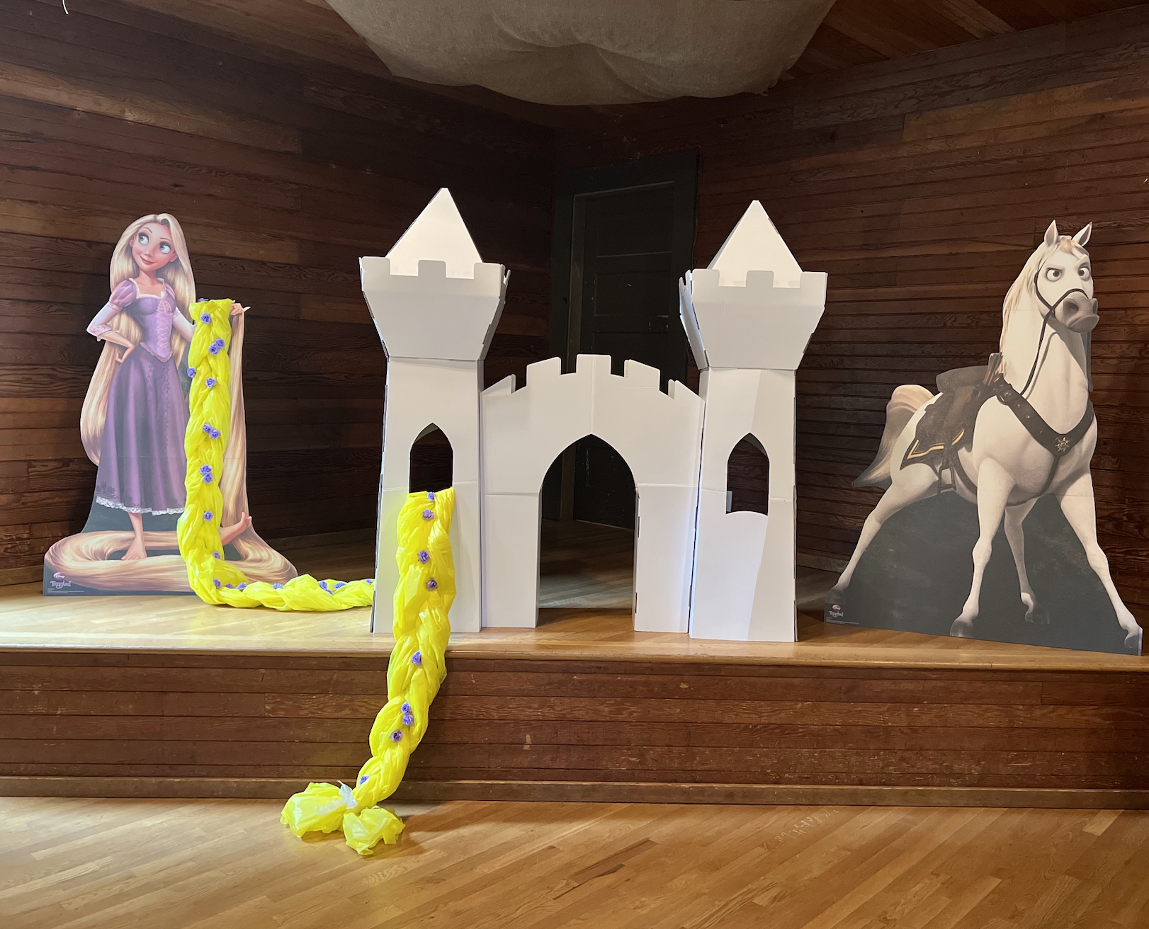 A cardboard castle with two towers on a wooden platform, flanked by cardboard cutouts of Rapunzel with long blonde hair holding a yellow braid decorated with purple flowers on the left and a white horse with a black bridle on the right.