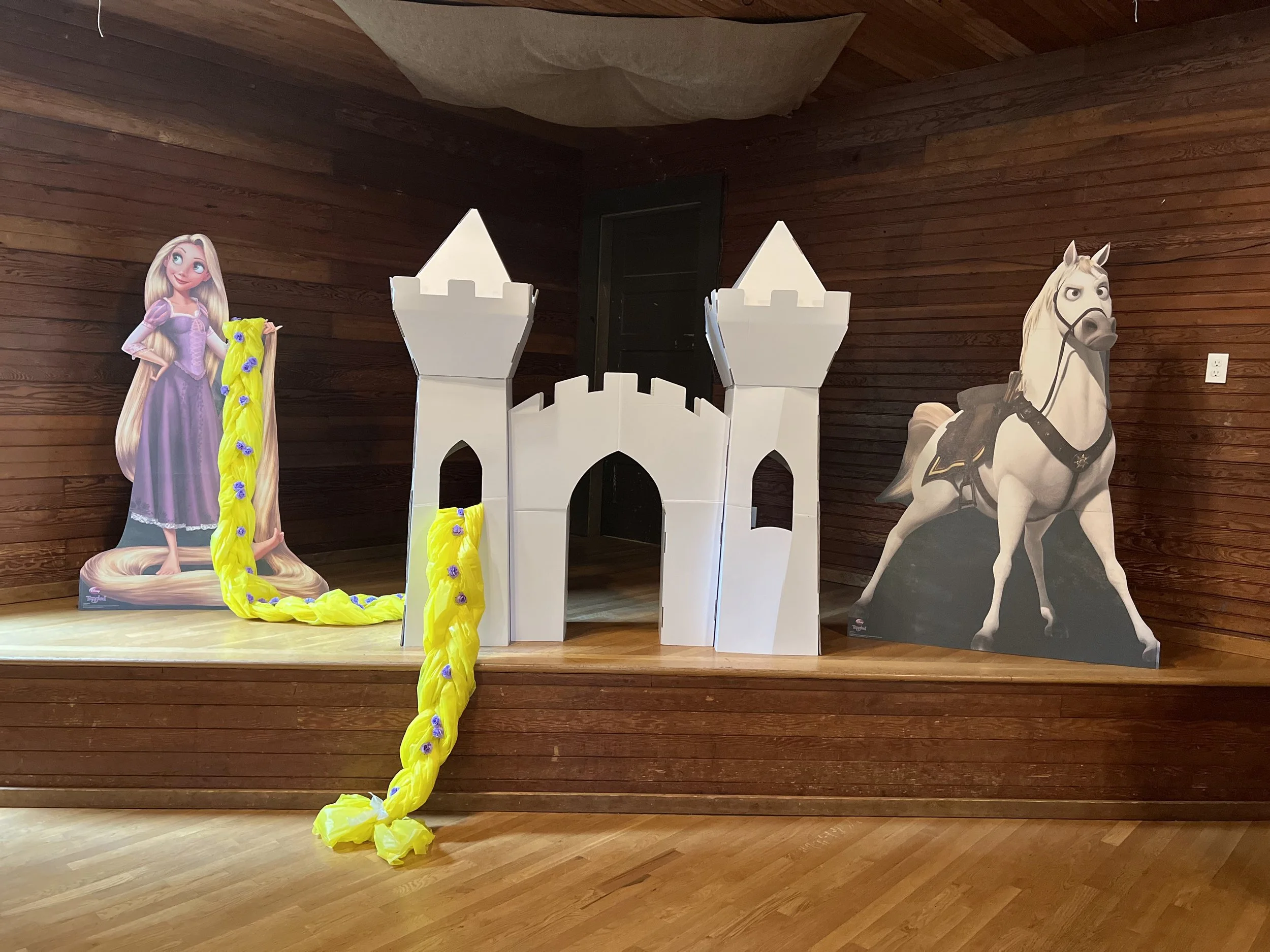 A children's play setup on a wooden stage featuring a cardboard castle, Rapunzel from Disney with long yellow braided hair adorned with purple flowers, and a white horse with a black harness. The castle is white with four towers, and the stage has a 