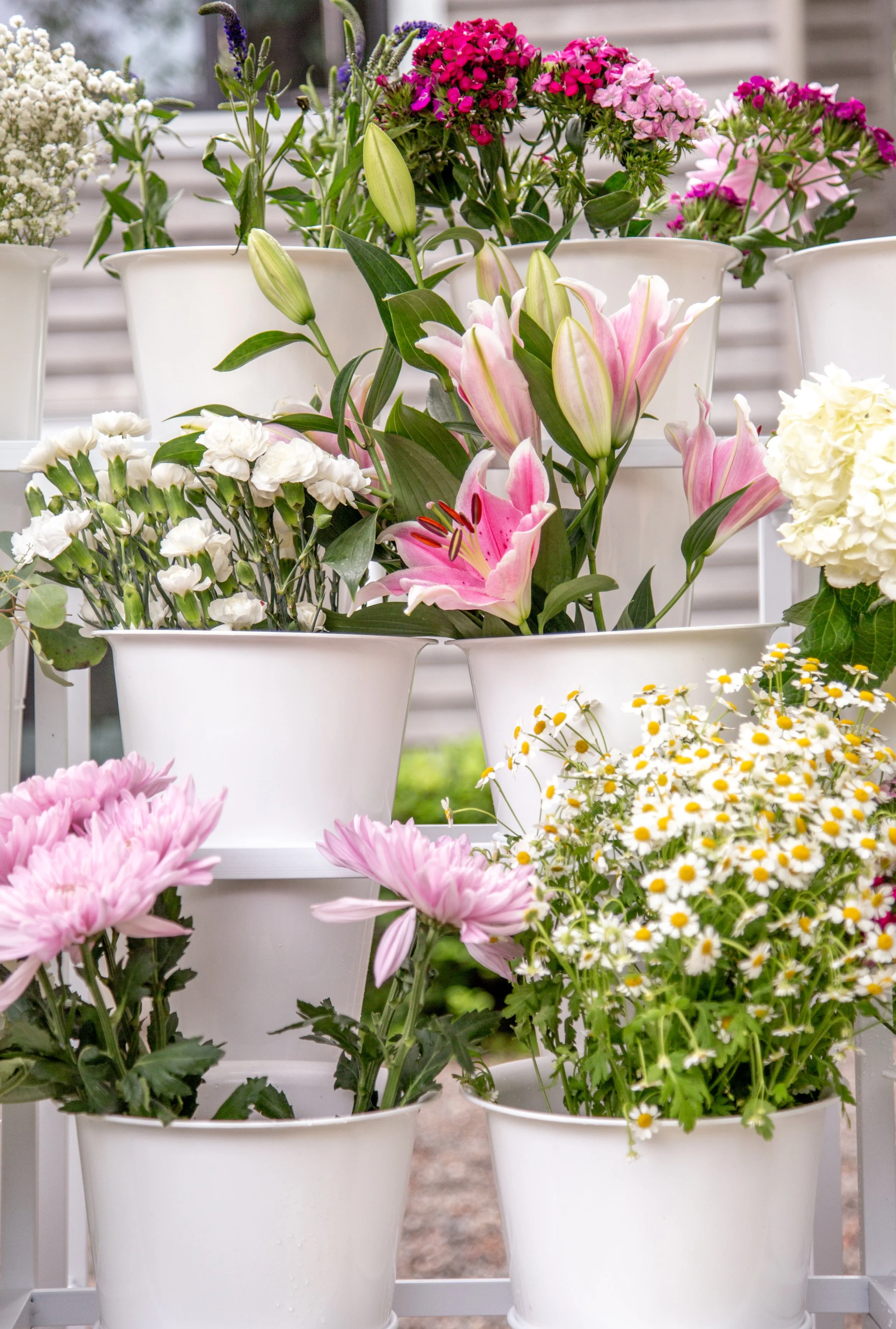 Various colorful flowers in white pots on a white stand, including pink lilies, white carnations, purple lavender, white daisies, and pink and purple alyssum, with a wooden building in the background.