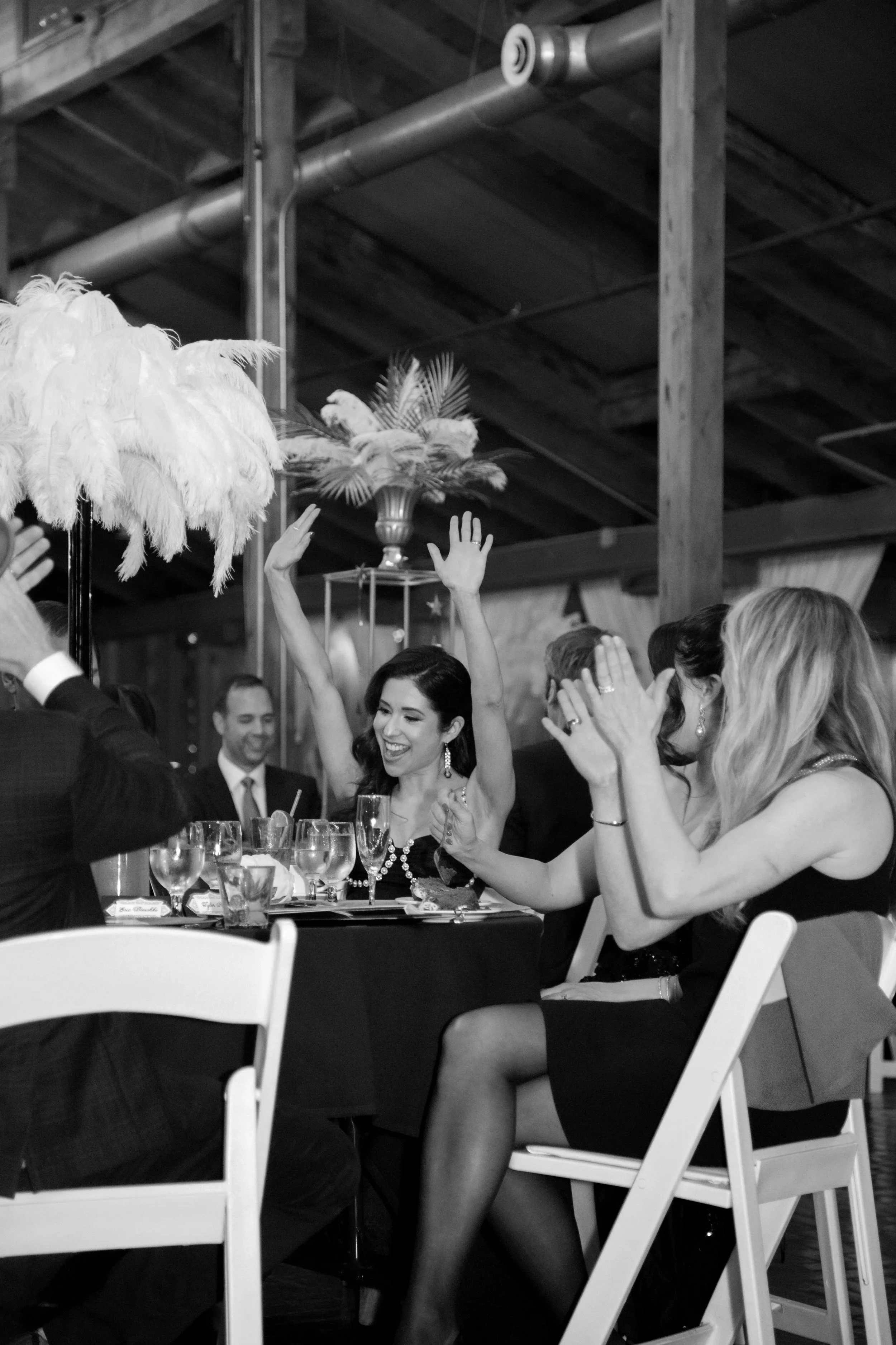 Women celebrating at a dinner event, clapping and raising hands, with glasses and table setting in front of them, in a rustic indoor venue.