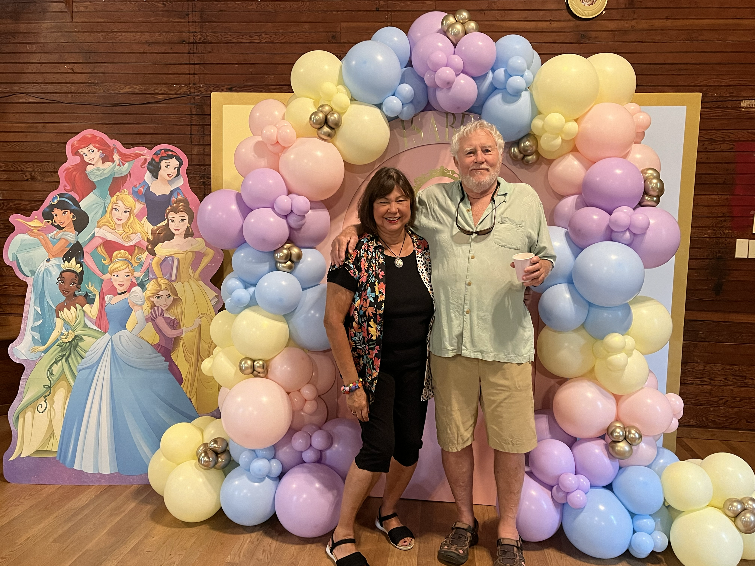Two people standing in front of a colorful balloon arch and a Disney princess cutout party decoration. The woman is wearing a black outfit with a floral kimono and the man is wearing a light green shirt and tan shorts, holding a drink.