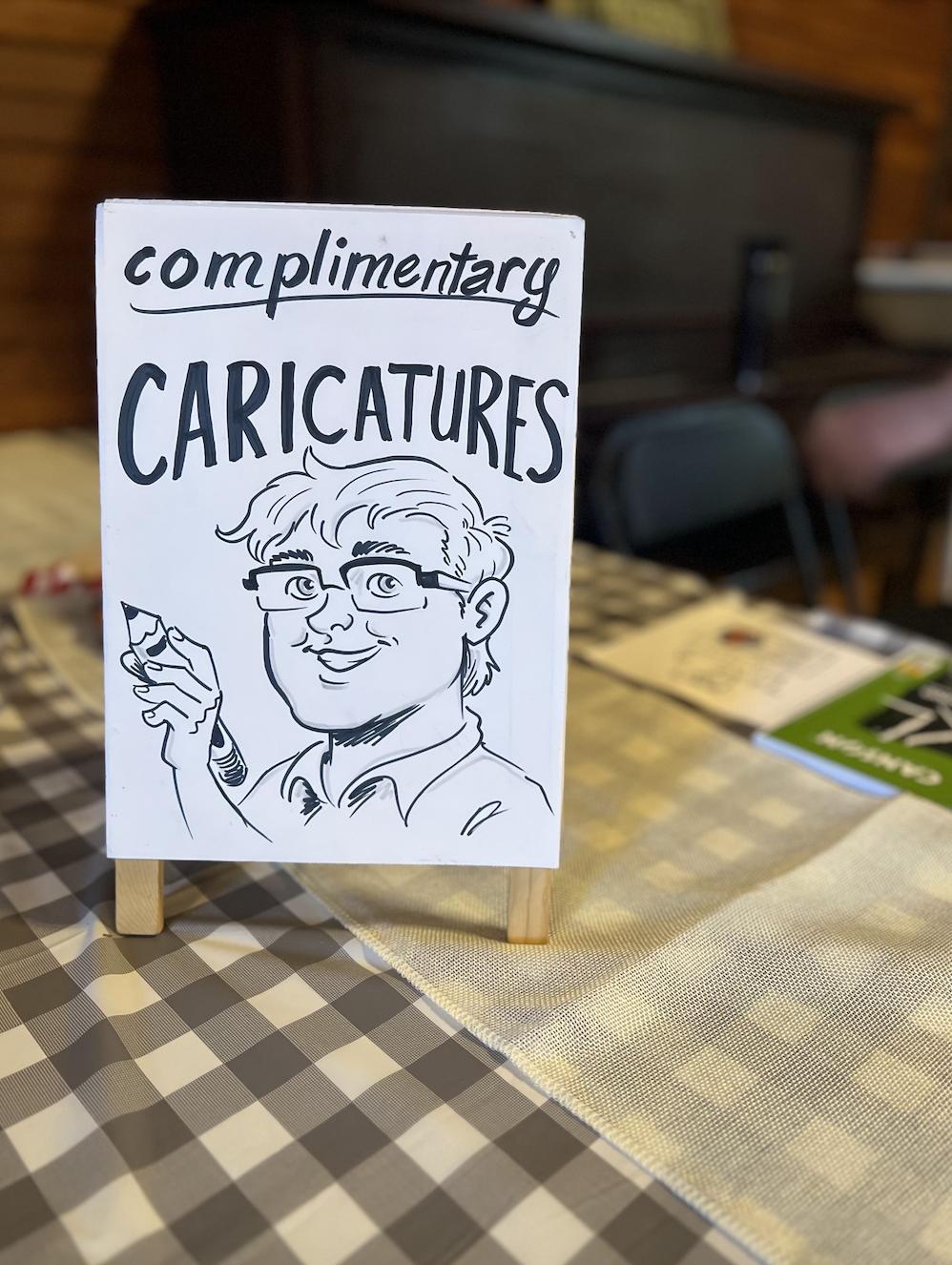 A small white sign on a table with a caricature drawing of a person holding a marker, advertising complimentary caricatures. The background shows a checkered tablecloth and some blurred papers and pamphlets.