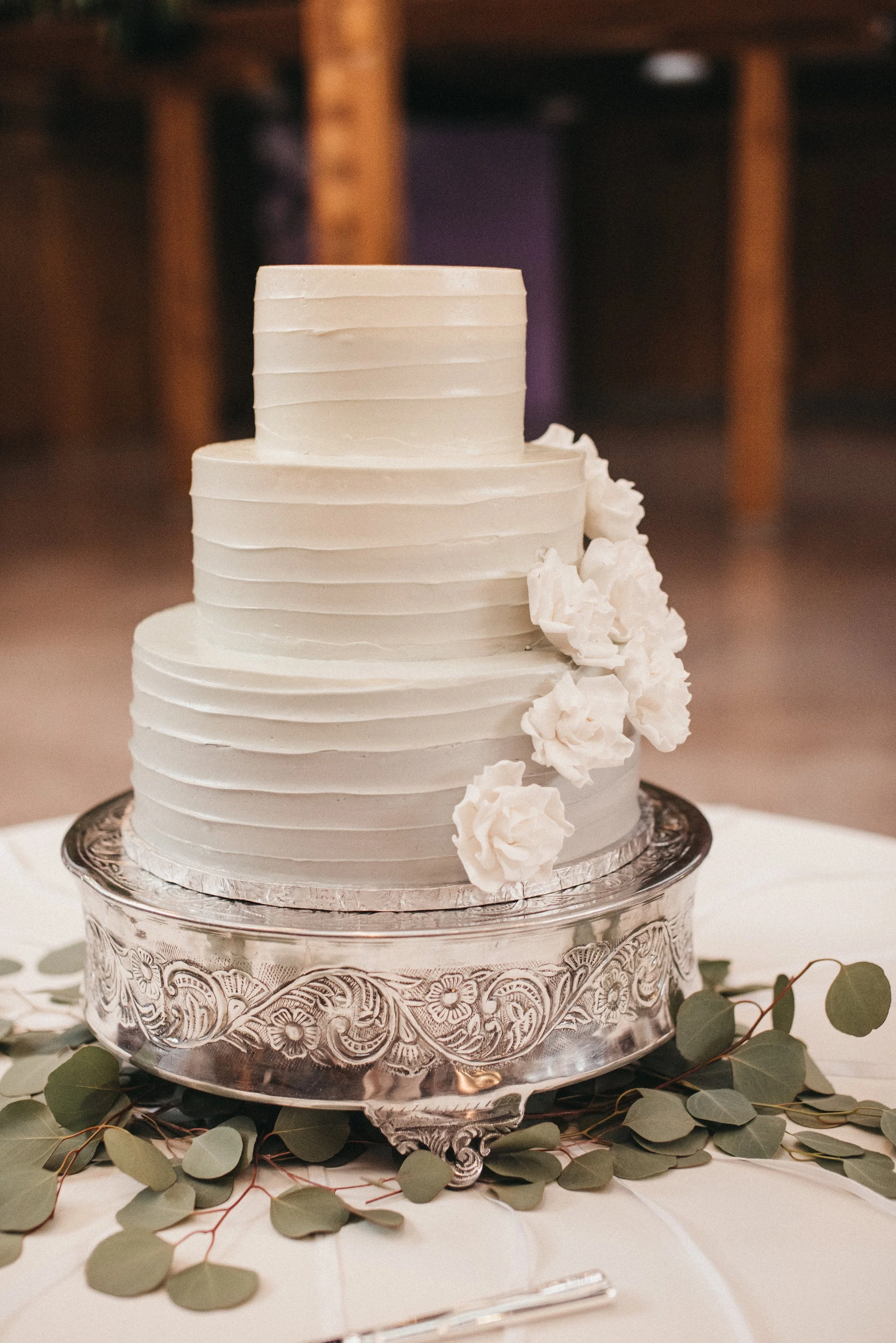 Three-tiered white wedding cake with horizontal ridges, decorated with white flowers, on an ornate silver cake stand surrounded by green leaves on a table.