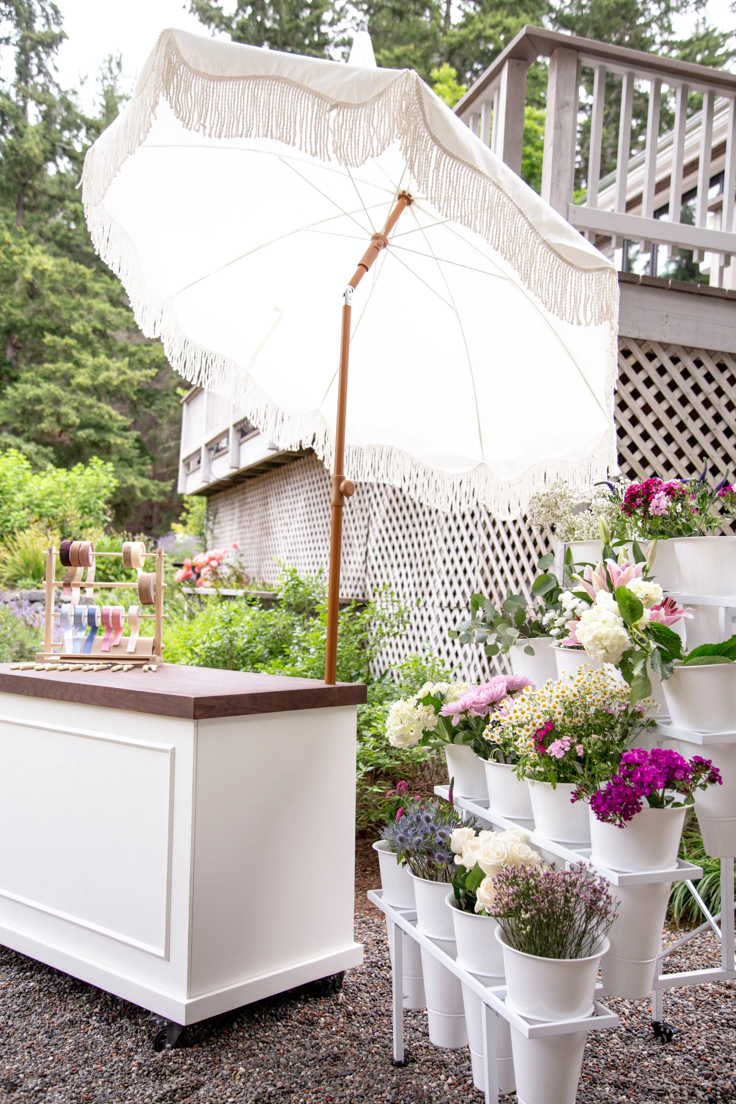 A white patio umbrella with fringe trim, surrounded by various colorful potted flowers on a tiered white flower stand, with a lattice fence, a deck, and greenery in the background.