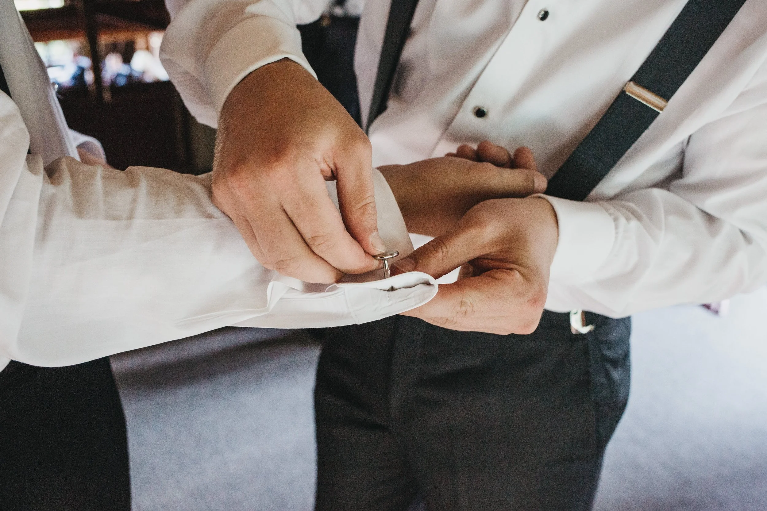 Two men in formal dress shirts and ties during a wedding, with one man pinning a cufflink onto the other's white shirt cuff.