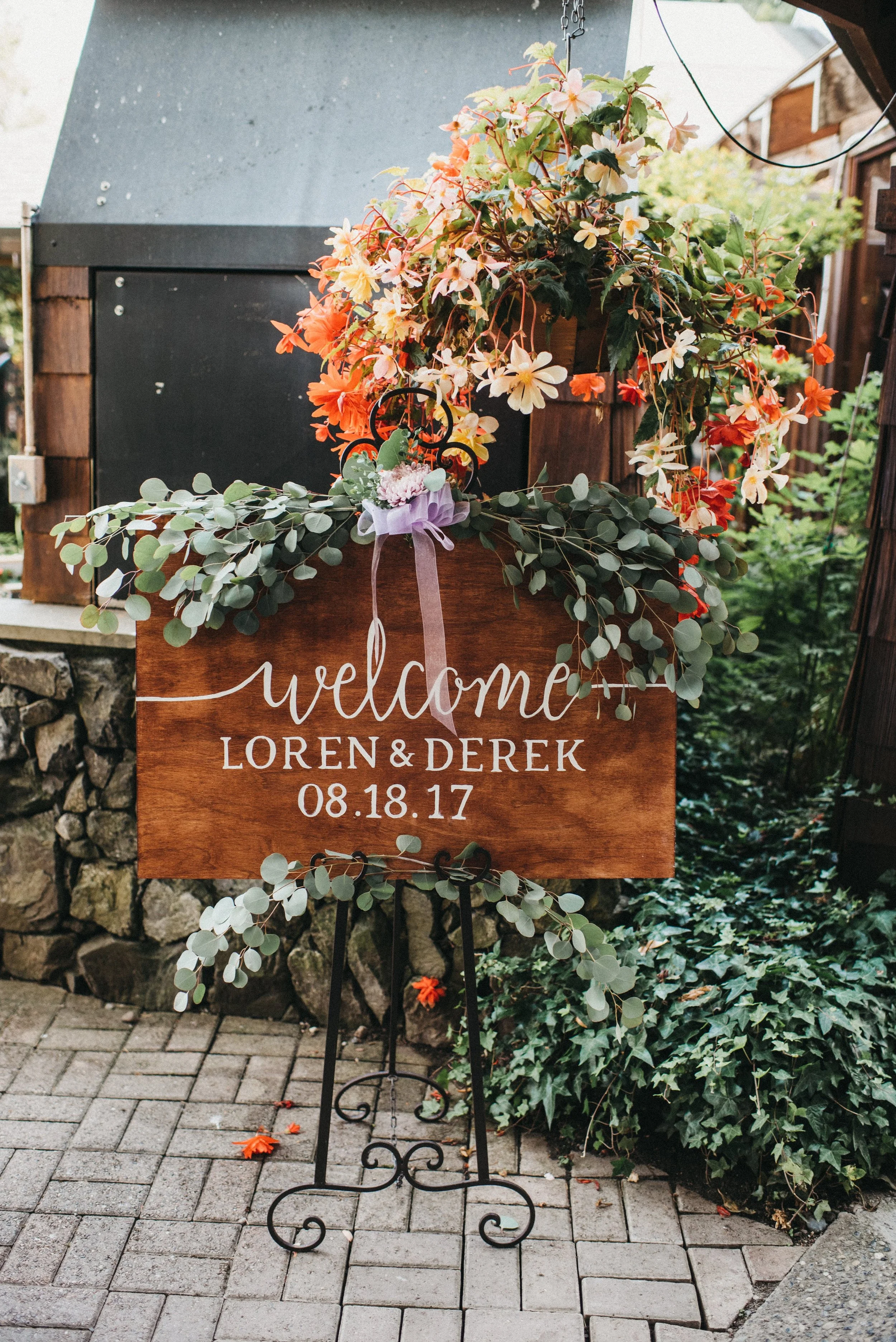 Wooden wedding welcome sign with flowers and greenery, displaying names Loren and Derek, and the date August 18, 2017.