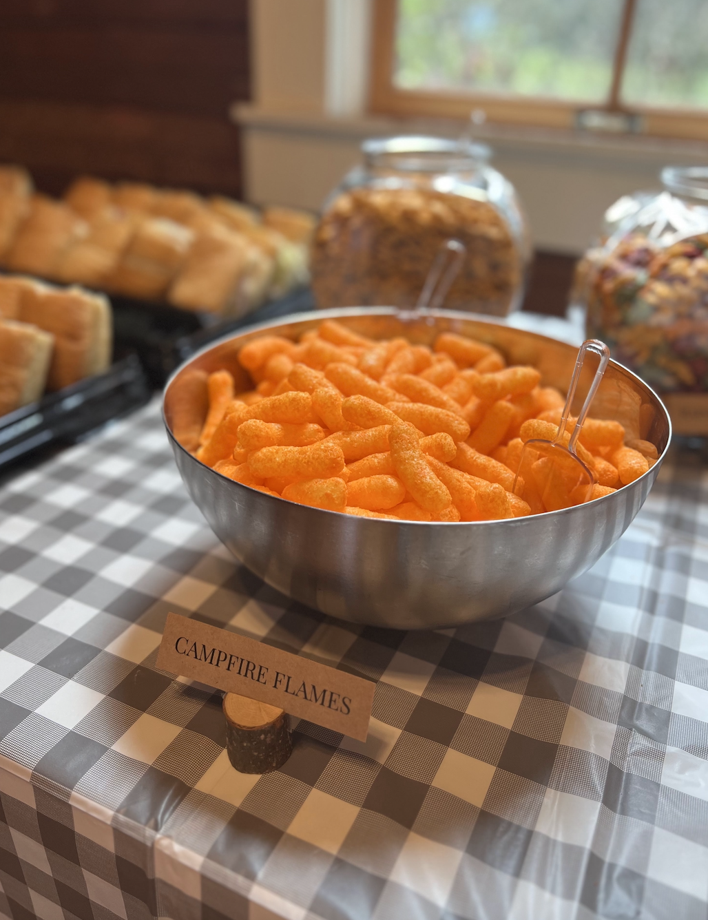 Bowl of orange cheese puffs labeled 'Campfire Flames' on a checkered tablecloth with bread and cereal jars in the background.