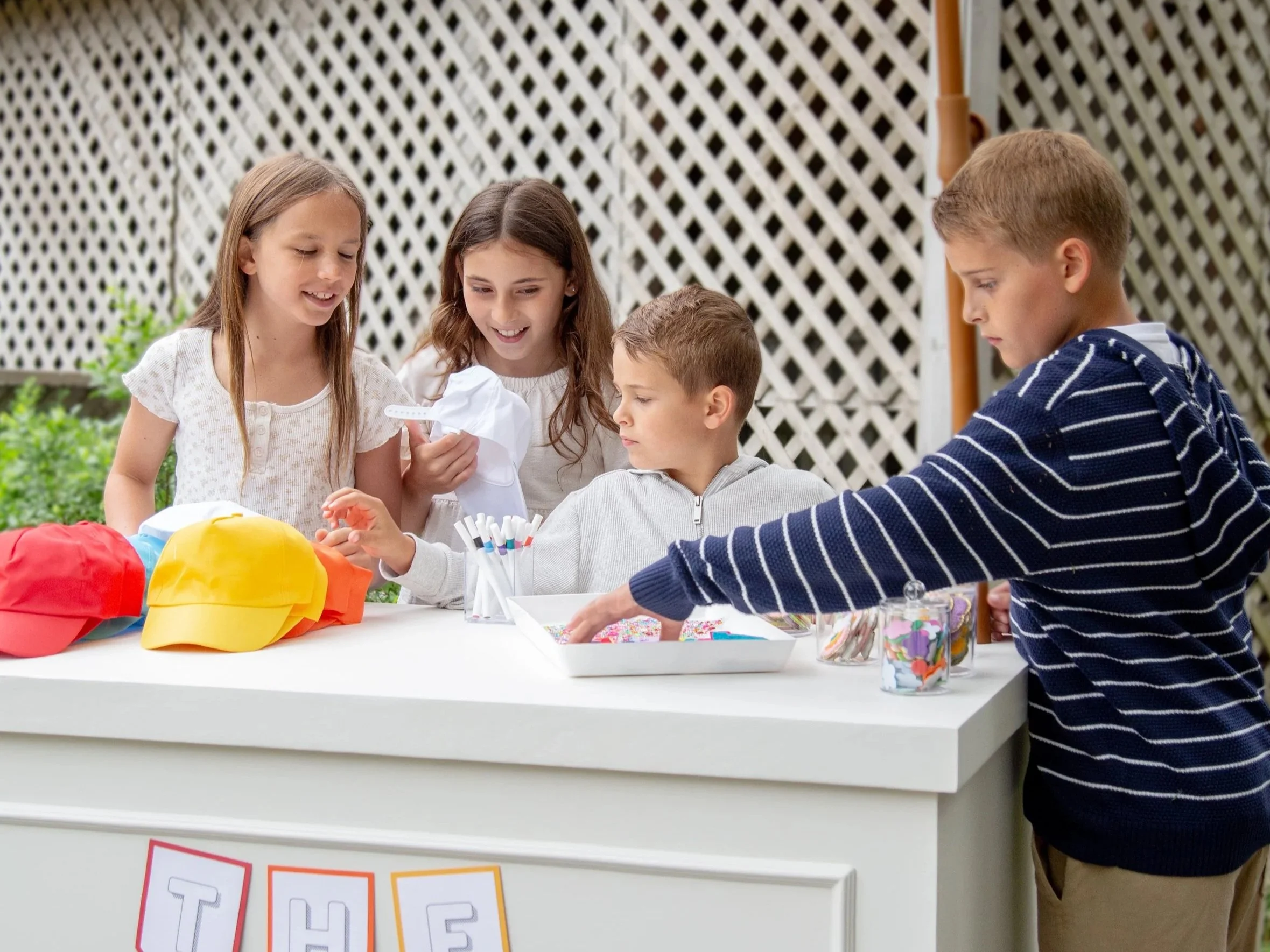 Four children are gathered around a table at an outdoor party, engaging with colorful party hats and supplies, with a lattice fence in the background.