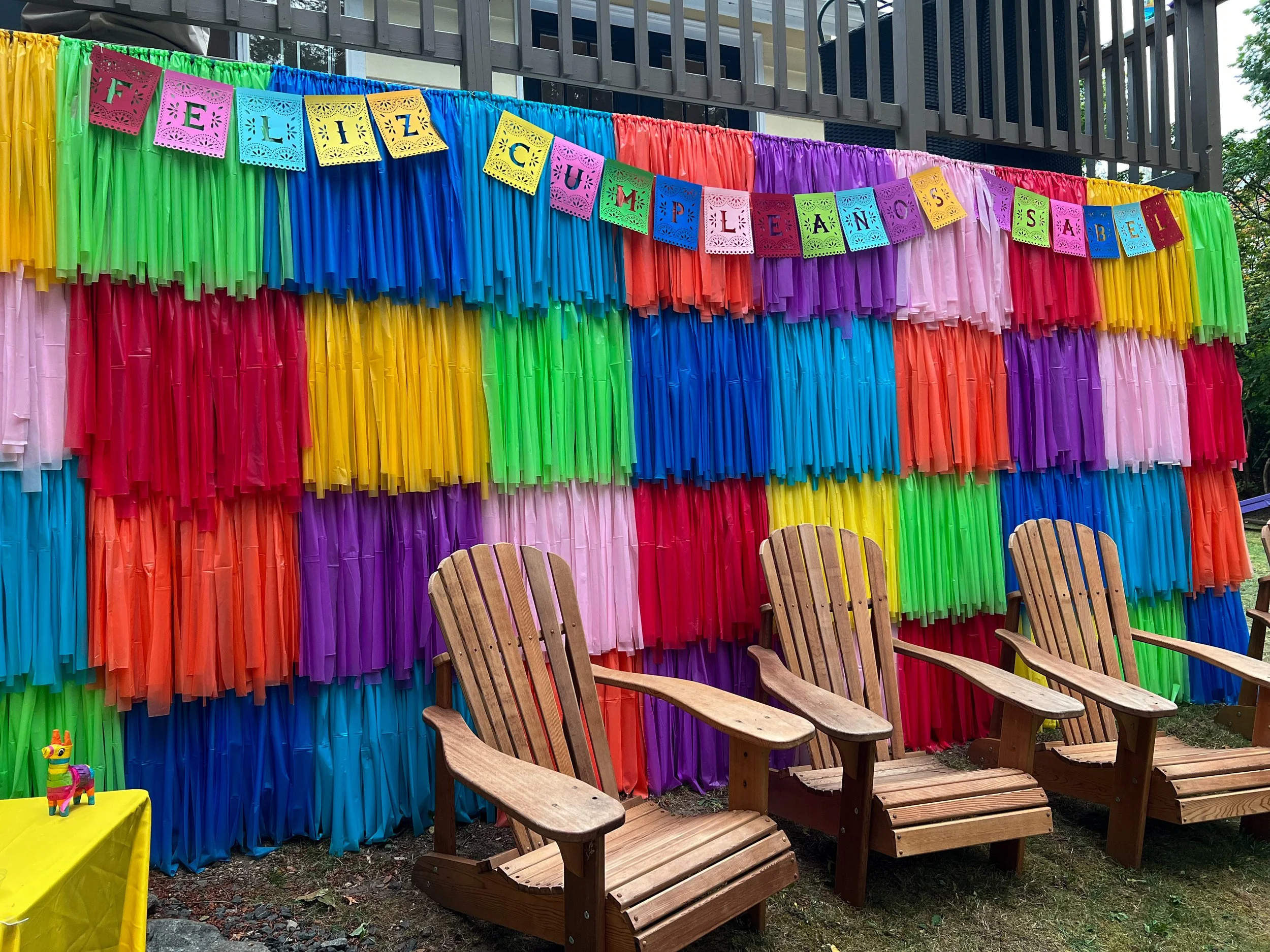 Rainbow-colored paper decorations hanging on a backdrop, with wooden Adirondack chairs in front and a small yellow table with a colorful llama figurine to the left. A festive banner at the top reads 'Ellic Cupcake and Sabel'.