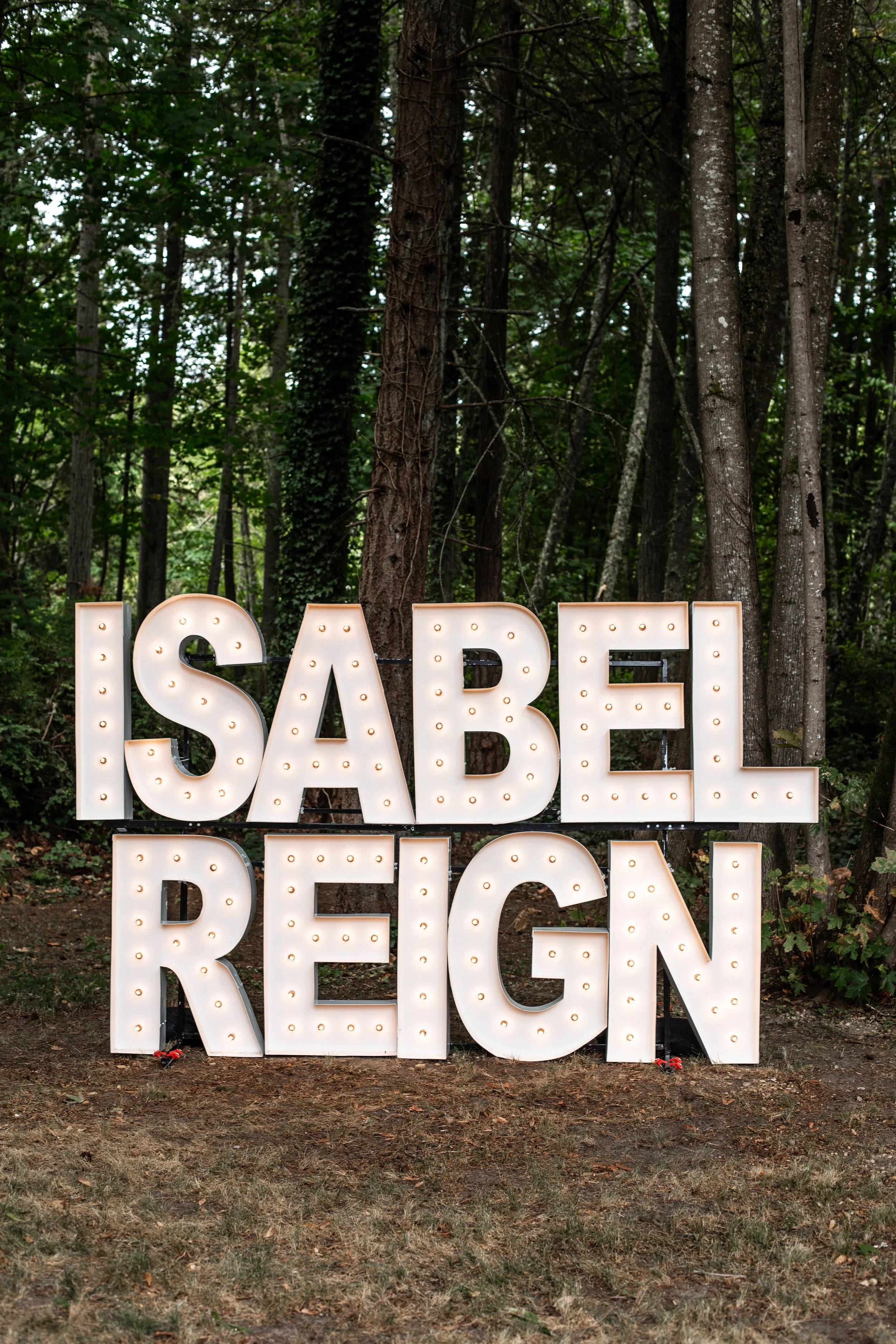 Large illuminated sign spelling 'ISABEL REIGN' set against a wooded outdoor background with trees and greenery.