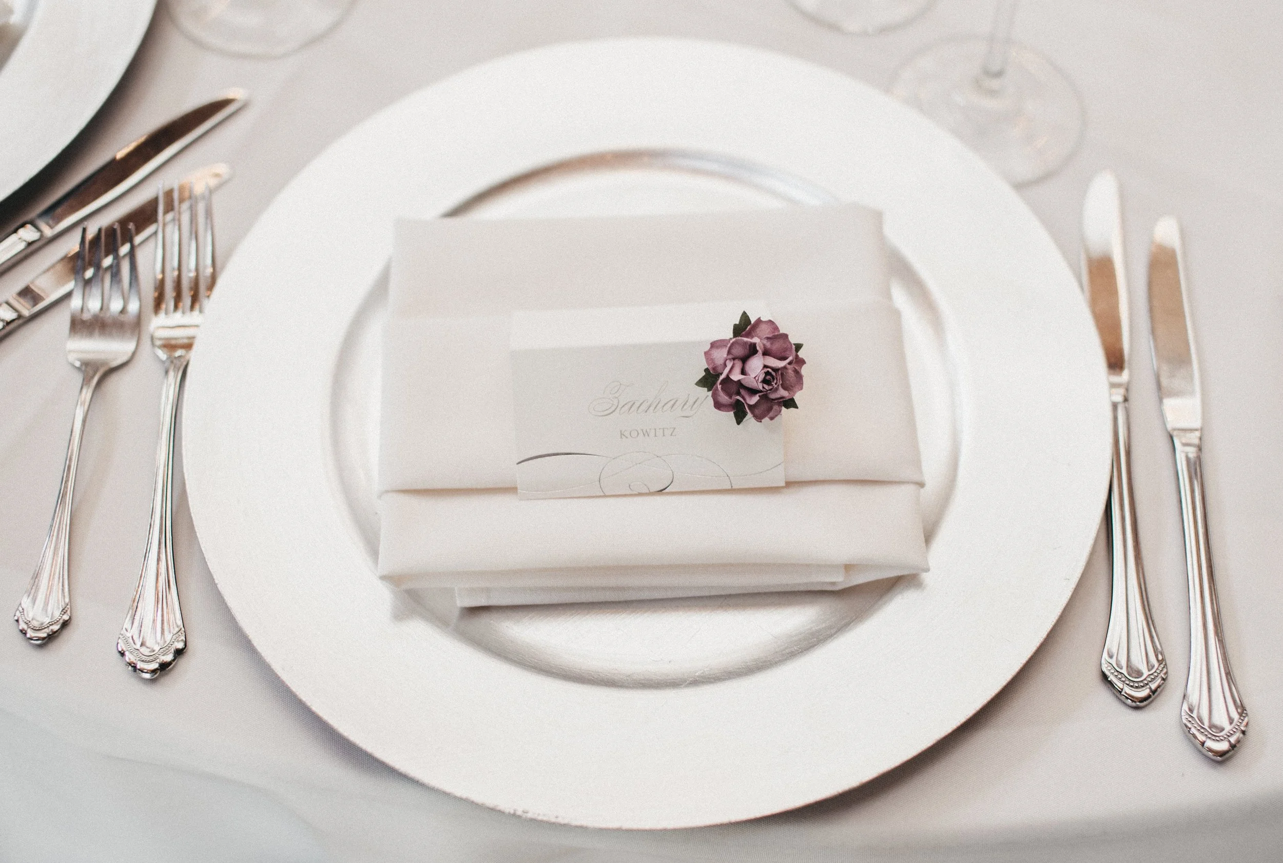 Elegant place setting with a white plate, a napkin, cutlery, and a small name card decorated with a purple flower, on a table with a white tablecloth, wine glasses in the background.