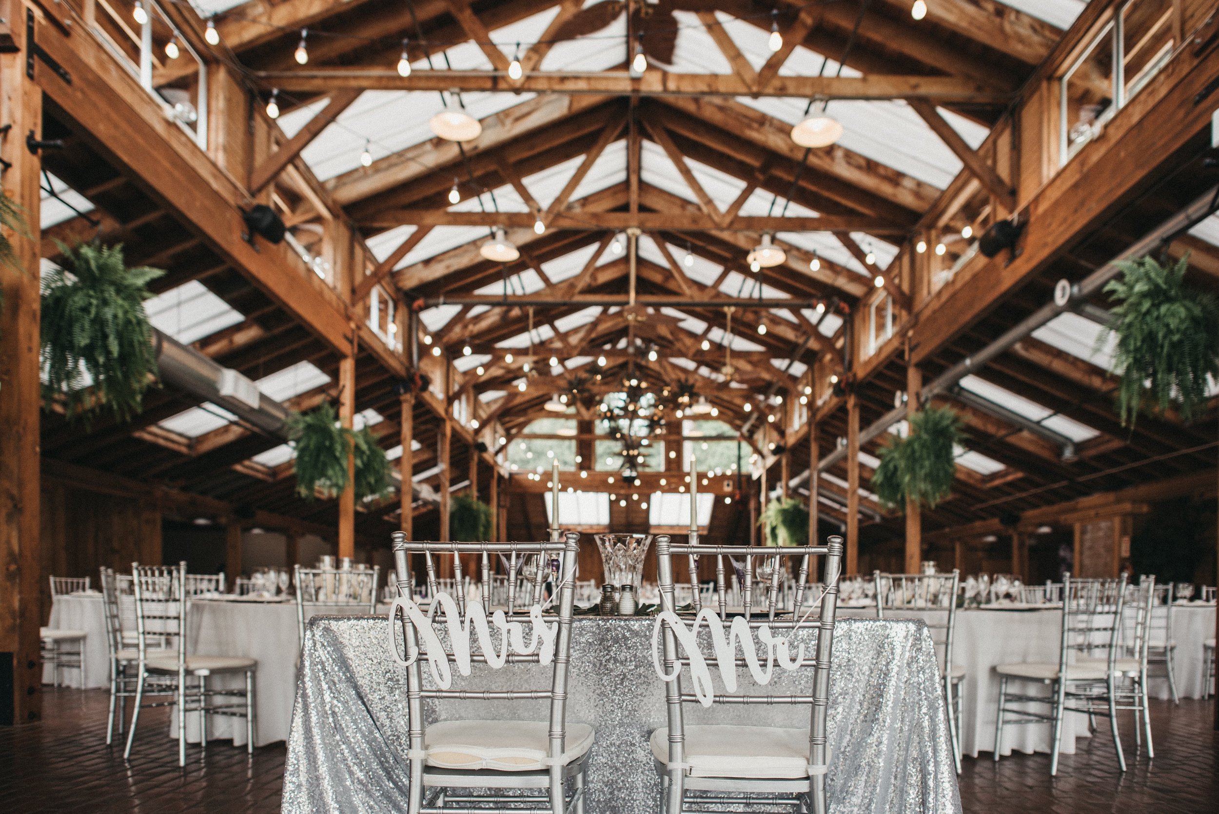 Elegant wedding reception in a rustic wooden venue with round tables, a silver sparkly tablecloth, tall candles, and greenery decorations hanging from the ceiling.