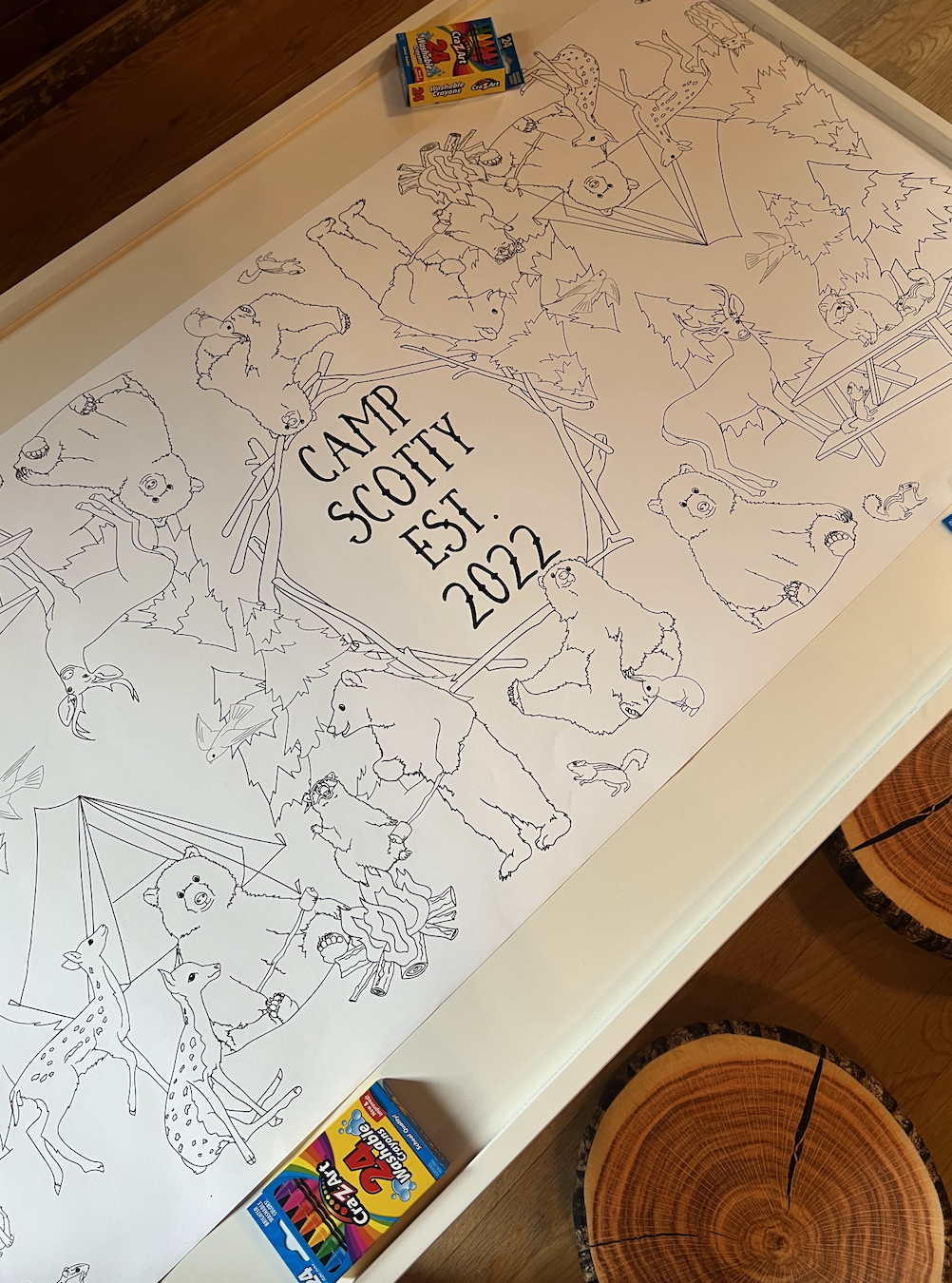 A large indoor activity sheet features outline drawings of various forest animals including bears, deer, and rabbits, with the text "CAMP SCOUTY EST. 2022" in the center. Crayons and colored markers are visible at the top of the sheet, and two packs 