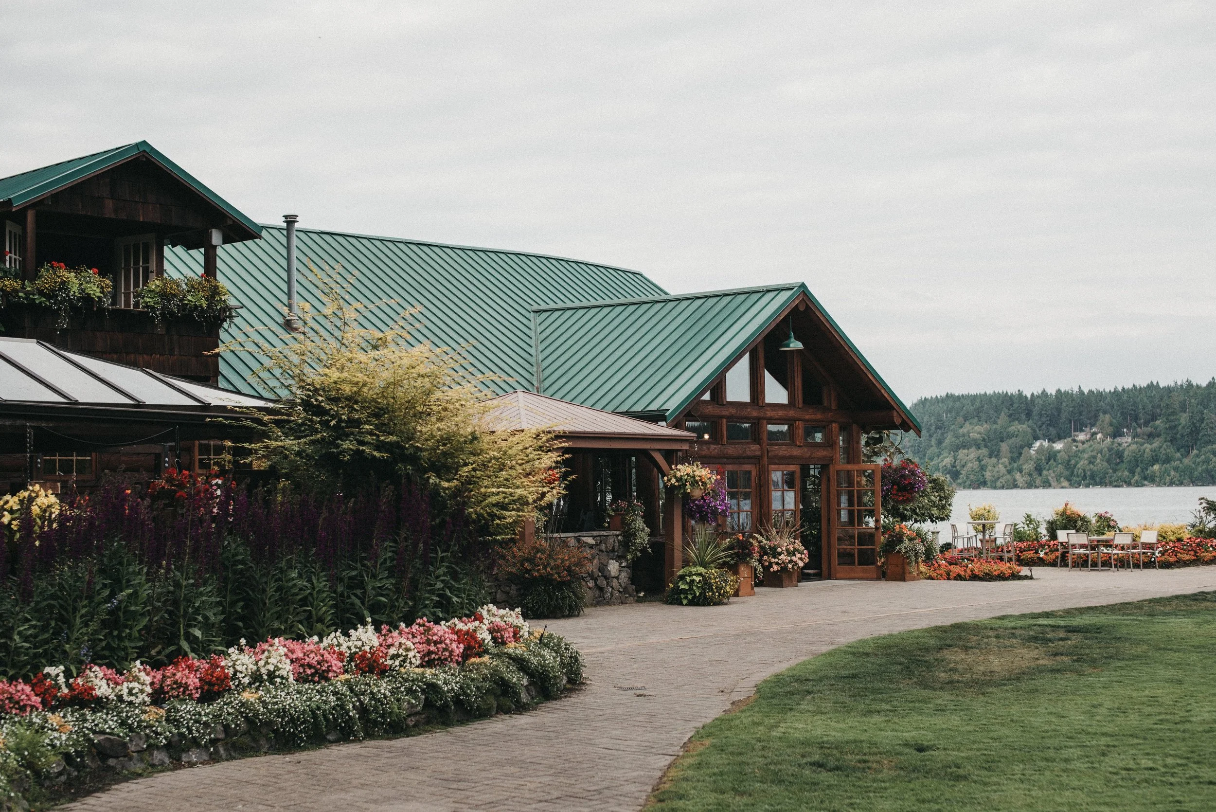 A lakeside building with a green metal roof, wooden accents, and large glass windows, surrounded by colorful flowers and plants, with outdoor seating and a lake in the background.