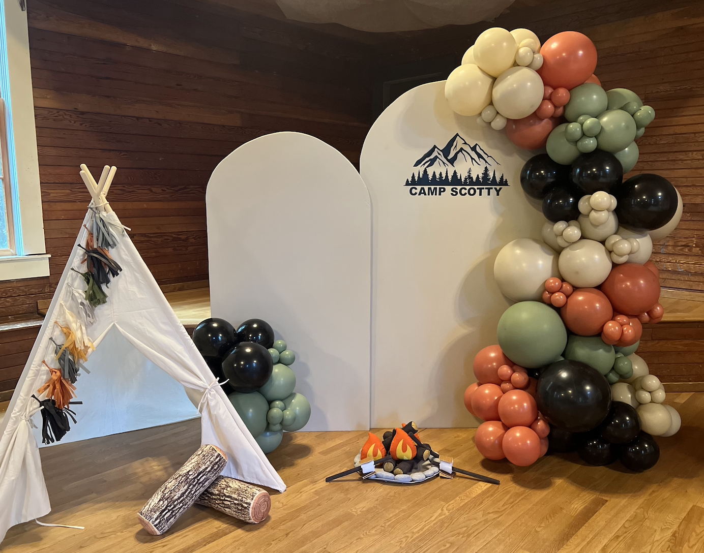 Camping-themed decoration with a white teepee, faux logs, a balloon arrangement of assorted colors, a white backdrop with 'Camp Scotty' and mountain graphic, and a campfire prop.