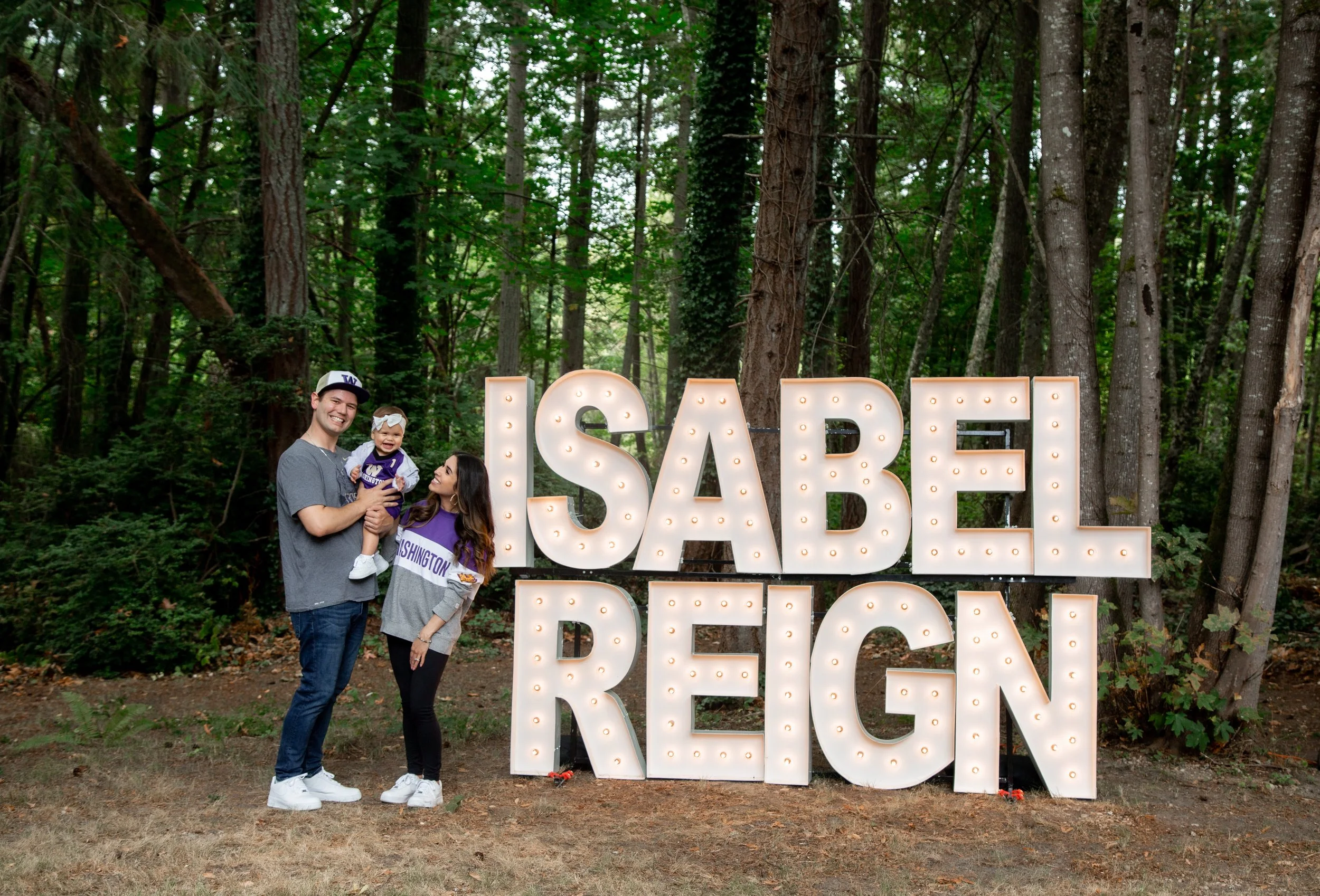 A family of three standing outdoors in front of large illuminated letters spelling 'SABEL REIGN' in a wooded area. The man is holding a baby girl, and the woman is standing beside them, all smiling.
