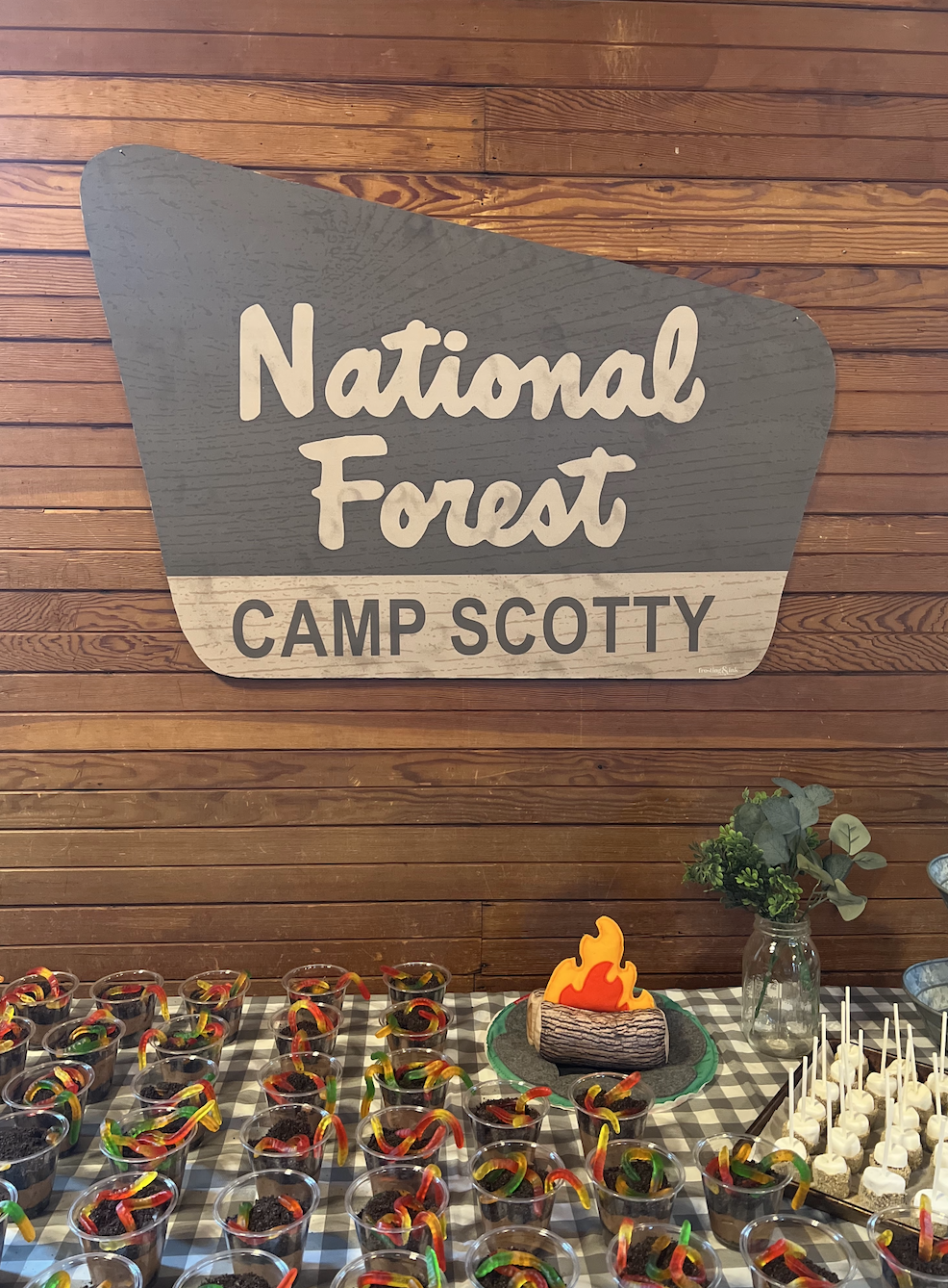 A table with Halloween-themed treats including small cups with worms and cookies, a decorative log with a flame, and a jar of greenery, against a wooden wall with a sign that reads 'National Forest Camp Scotty'.