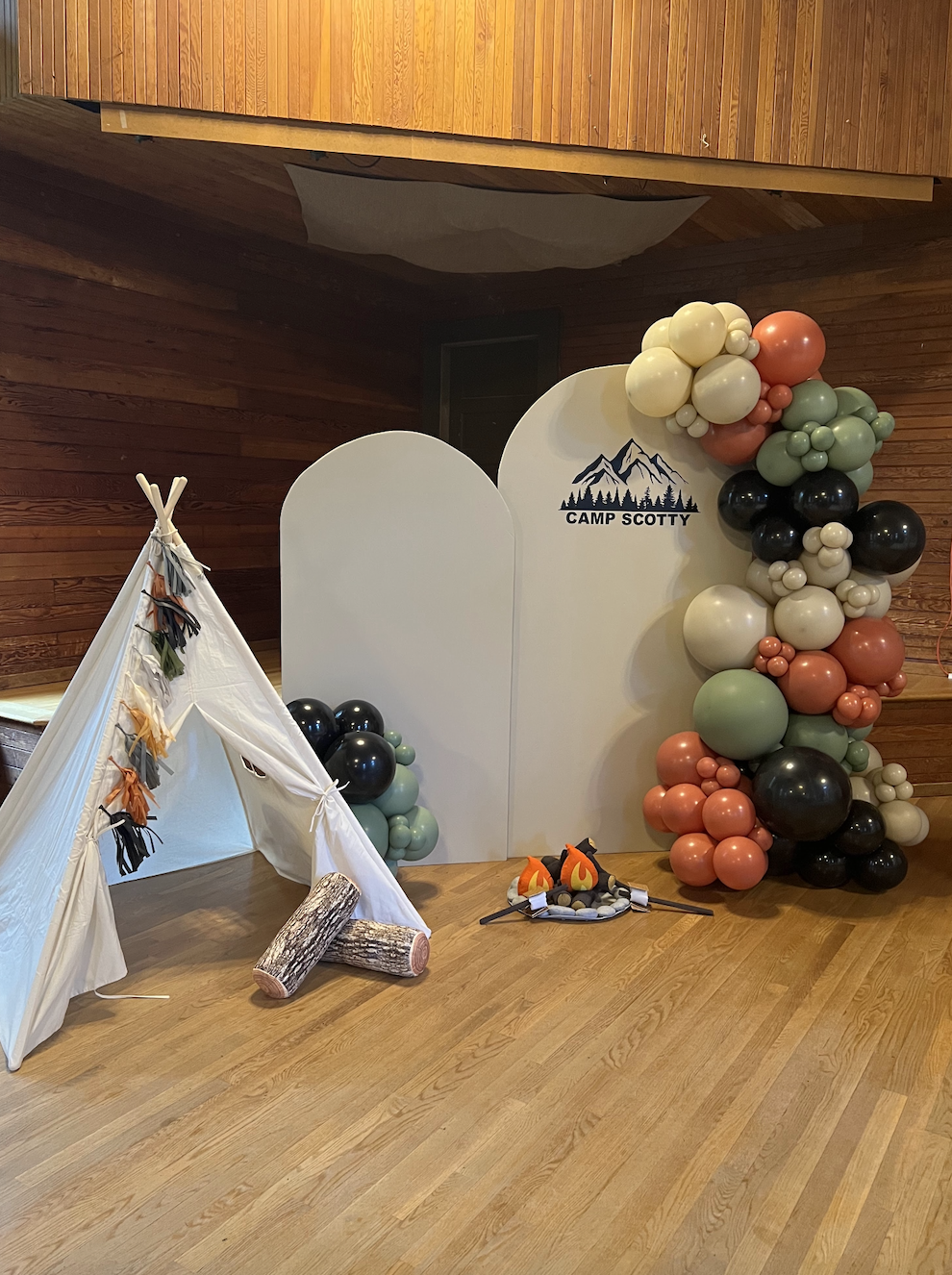 Decorative camping-themed display with a white teepee, artificial logs, and balloons shaped like mountains. The backdrop features a mountain graphic with the words 'Camp Scotty' and a balloon arrangement resembling a mountain landscape in black, peac